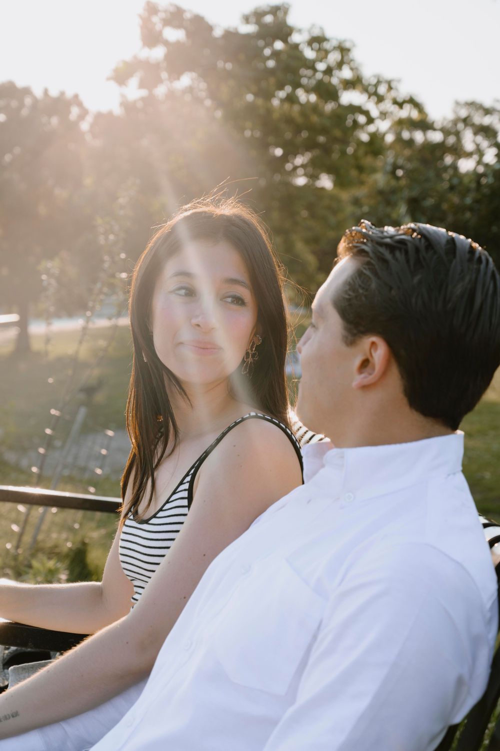 A romantic couple sits together in a park during golden hour with sun flares in the background.