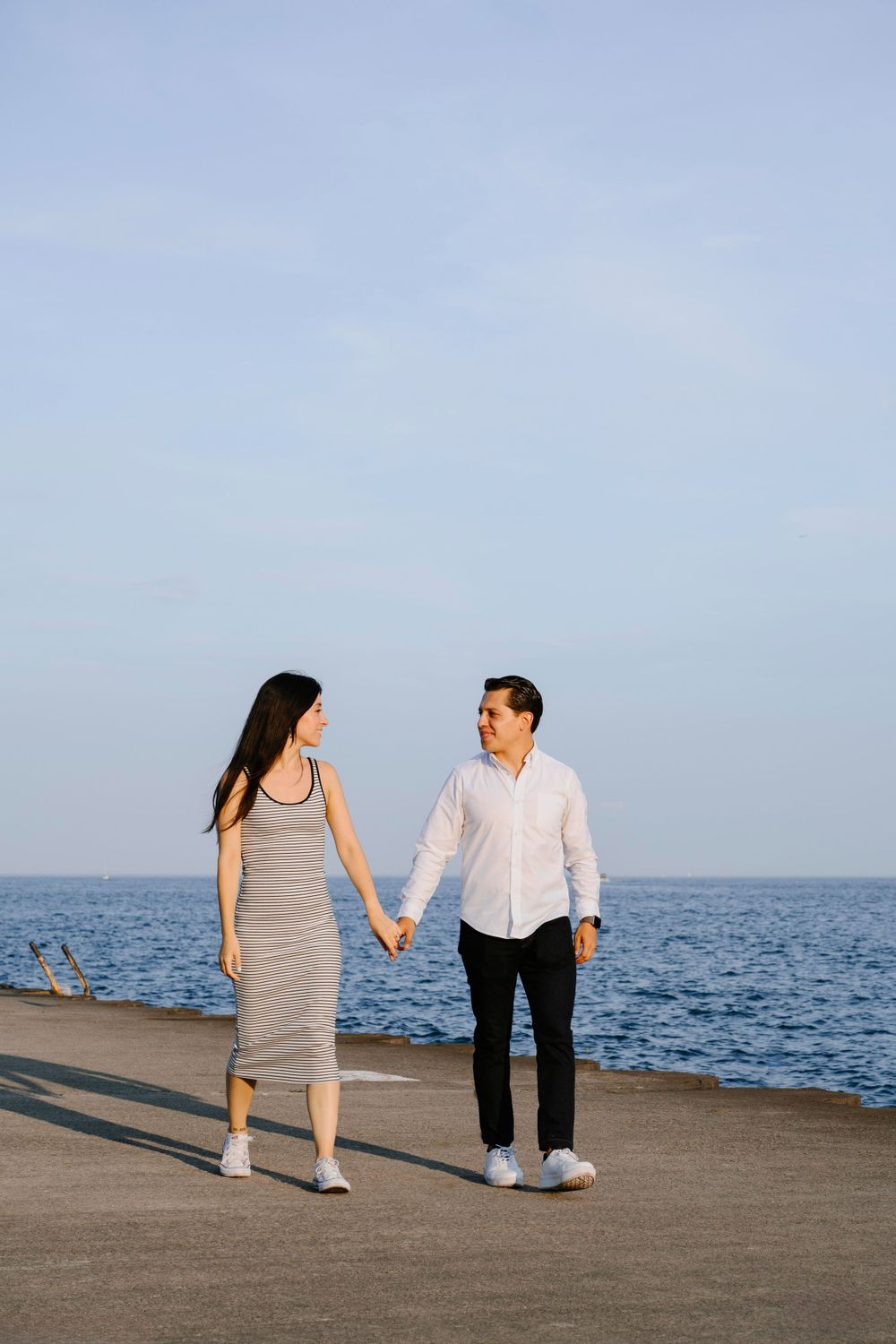 A romantic couple walking hand in hand along a seaside pier at sunset with the ocean in the background.