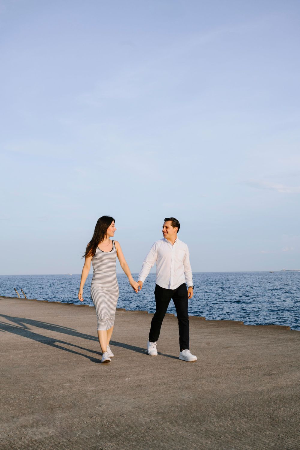 A couple holding hands while walking along a waterfront pier with ocean views.
