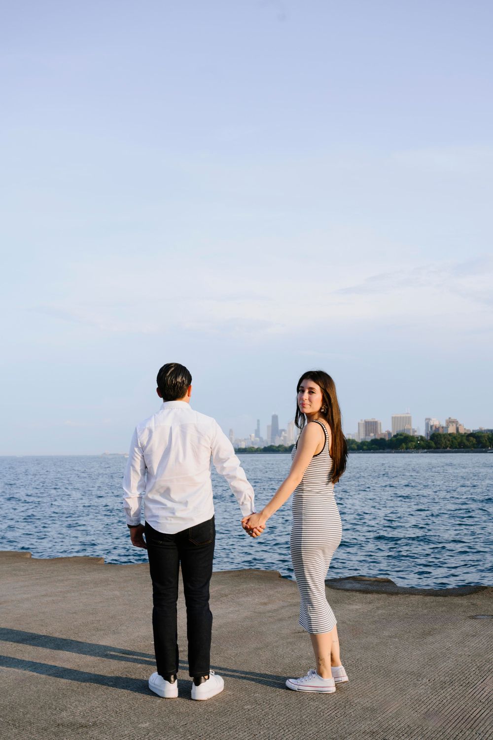 Couple holds hands while standing on a pier with city skyline in the background.