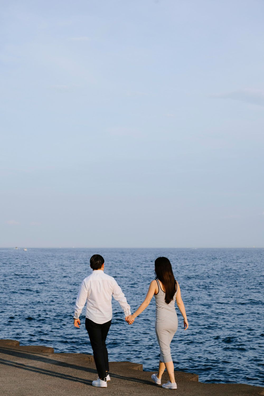 Two people walking hand in hand on a concrete path along the ocean on a bright clear day.
