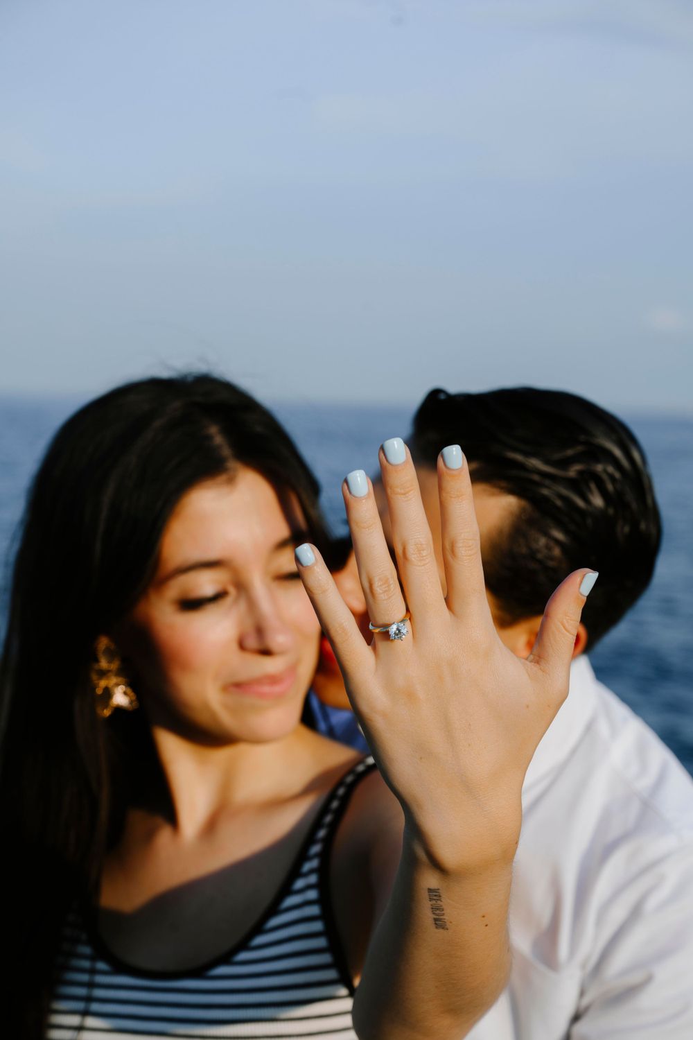 A newly engaged couple shares an intimate moment as she shows off her engagement ring by the ocean.