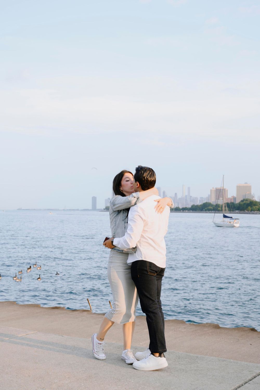 A couple embraces while standing on a waterfront walkway with a city skyline visible in the background.