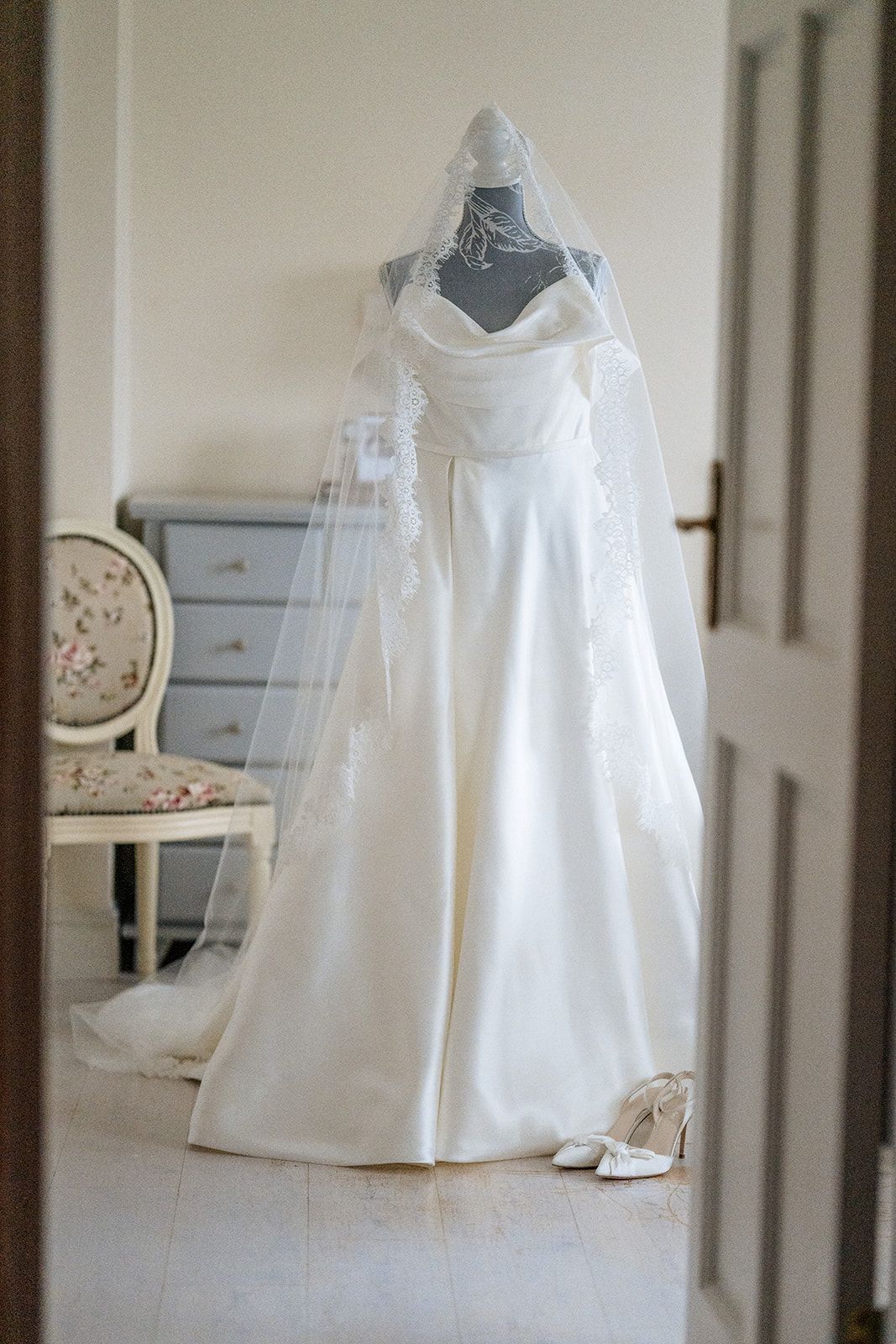 White wedding gown with veil displayed on a dress form in an elegant room with vintage furniture and white decor.