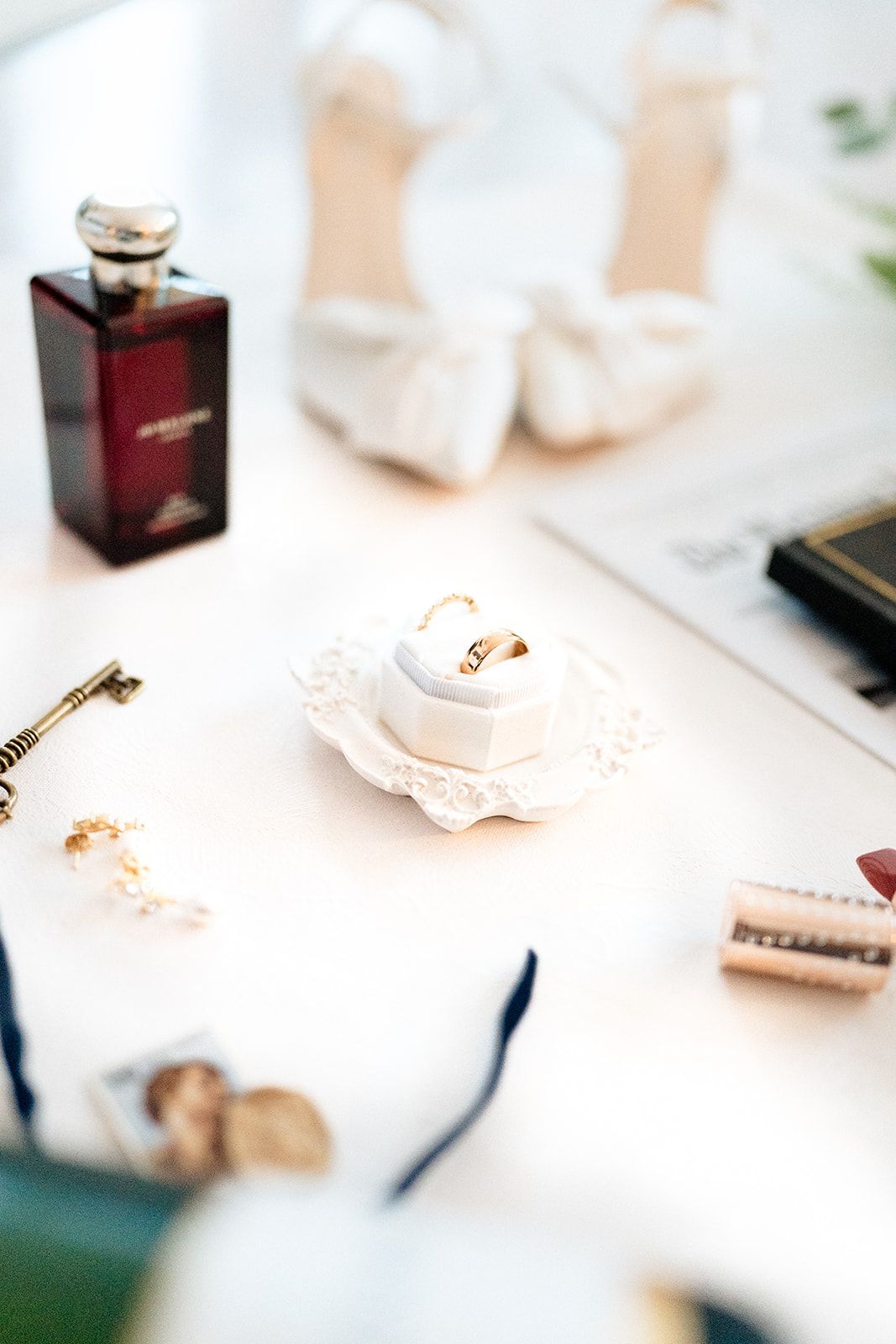 Wedding accessories including a vintage perfume bottle, delicate ring holder, and white bridal shoes arranged on a light surface.
