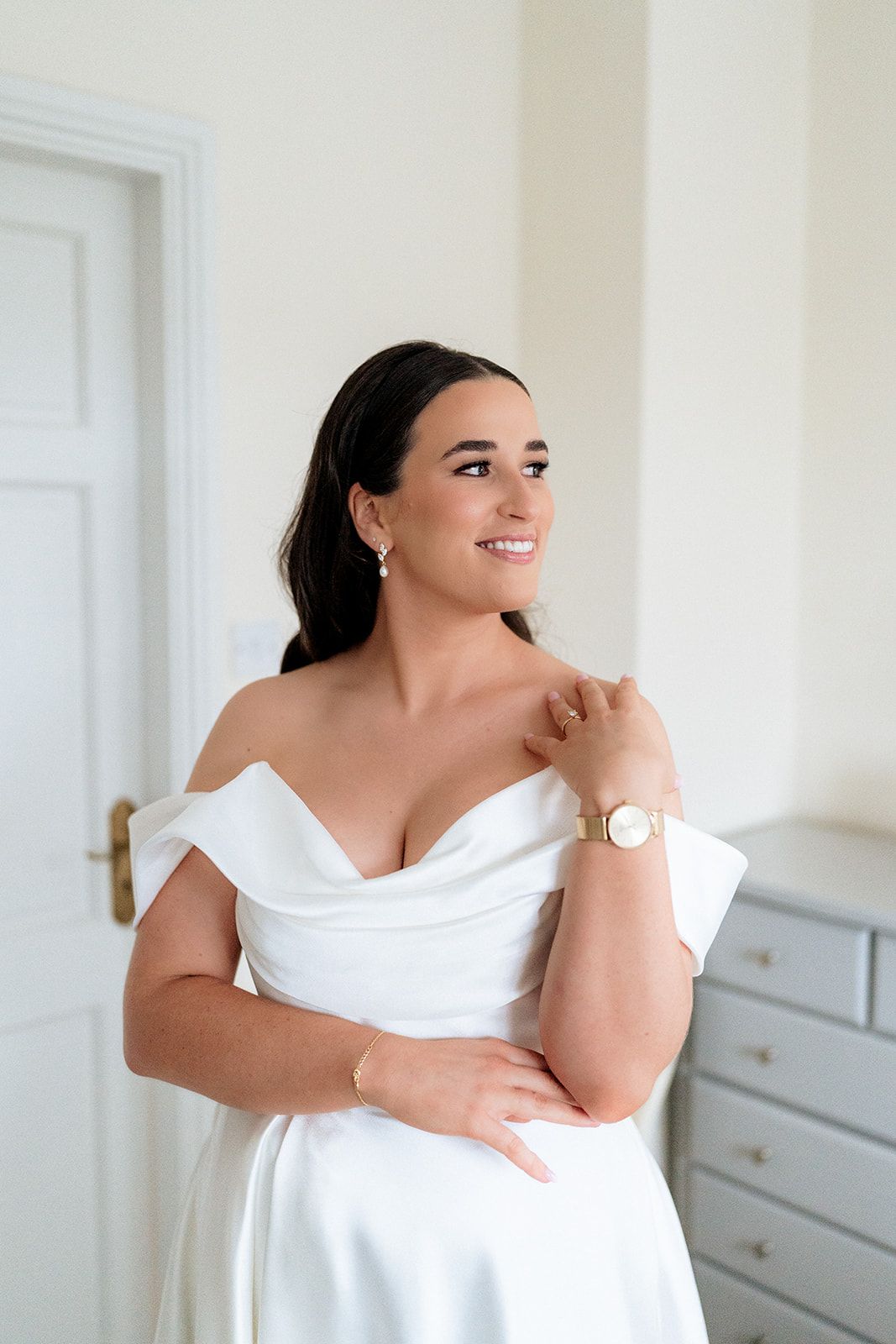 A bride wearing an elegant off-shoulder white wedding dress smiles while standing in a bright room with white walls.