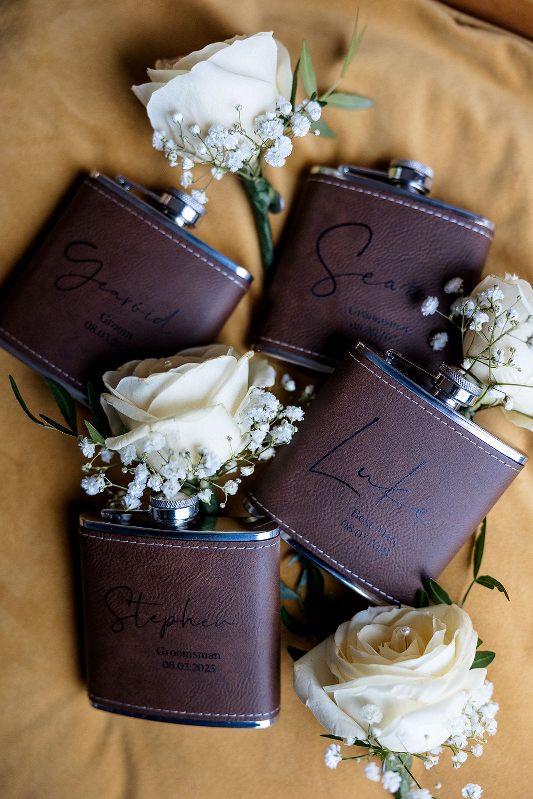 Four brown leather flasks surrounded by white roses and baby's breath flowers arranged on tan fabric backdrop.