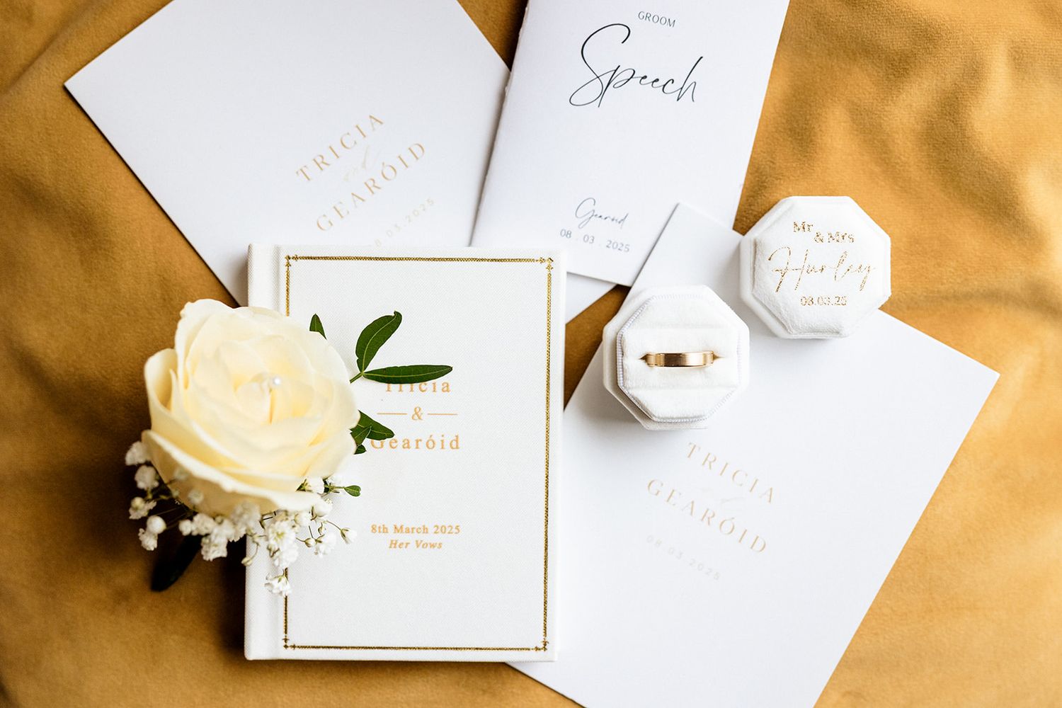 Elegant white wedding stationery set with cream rose and gold accents arranged on a golden fabric backdrop.