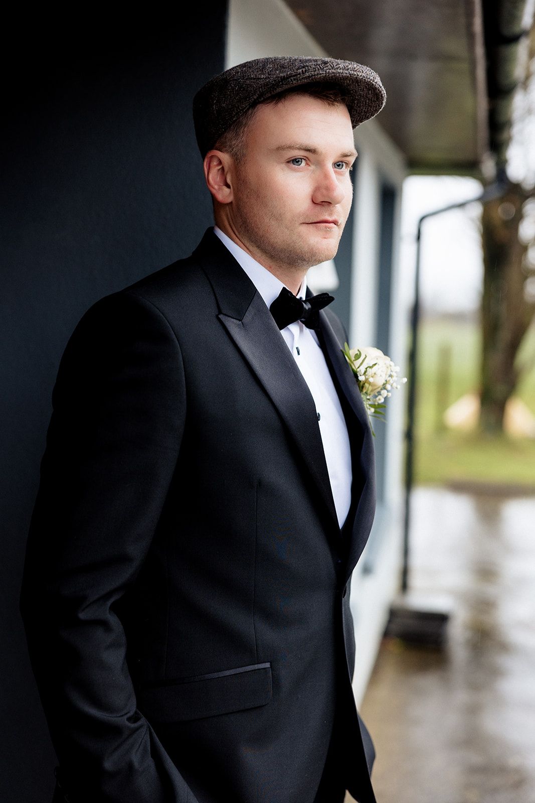 Groom in classic black tuxedo and brown flat cap poses against dark wall during formal wedding portraits.