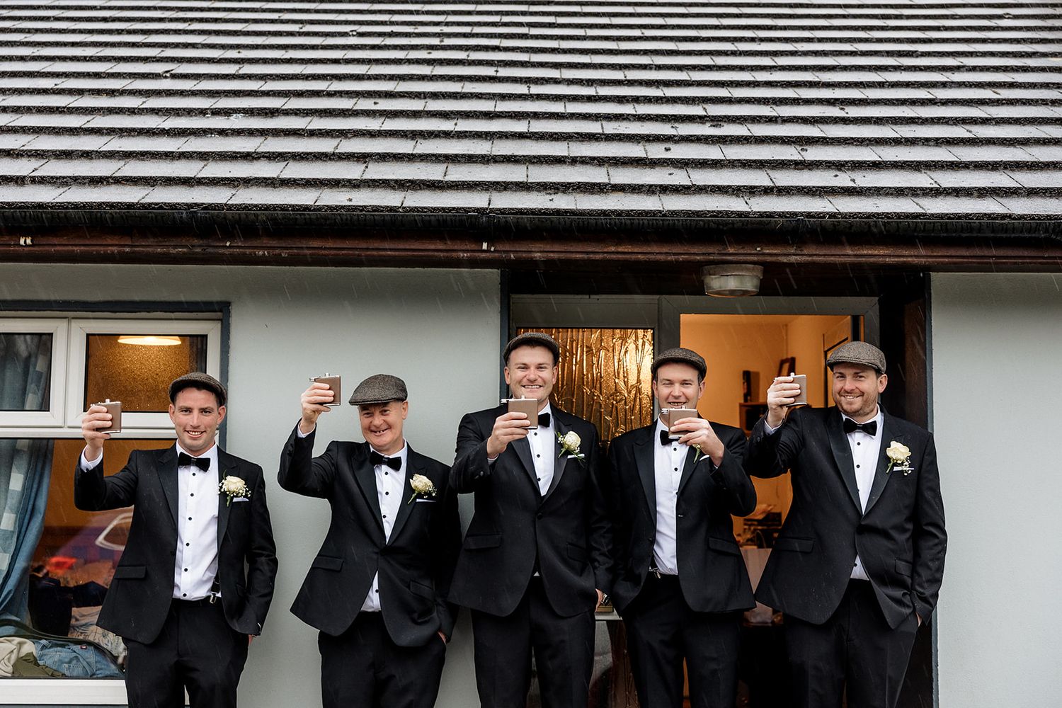 Five groomsmen in black tuxedos and bow ties stand outside a building entrance with warm lighting.