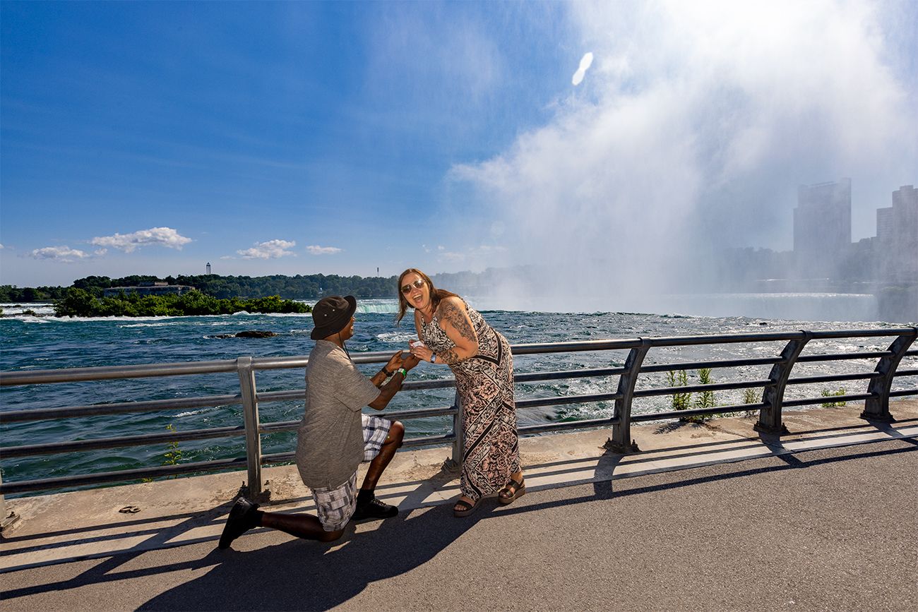 Two figures stand by the railing on a walkway near Niagara Falls with mist and blue sky in the background.