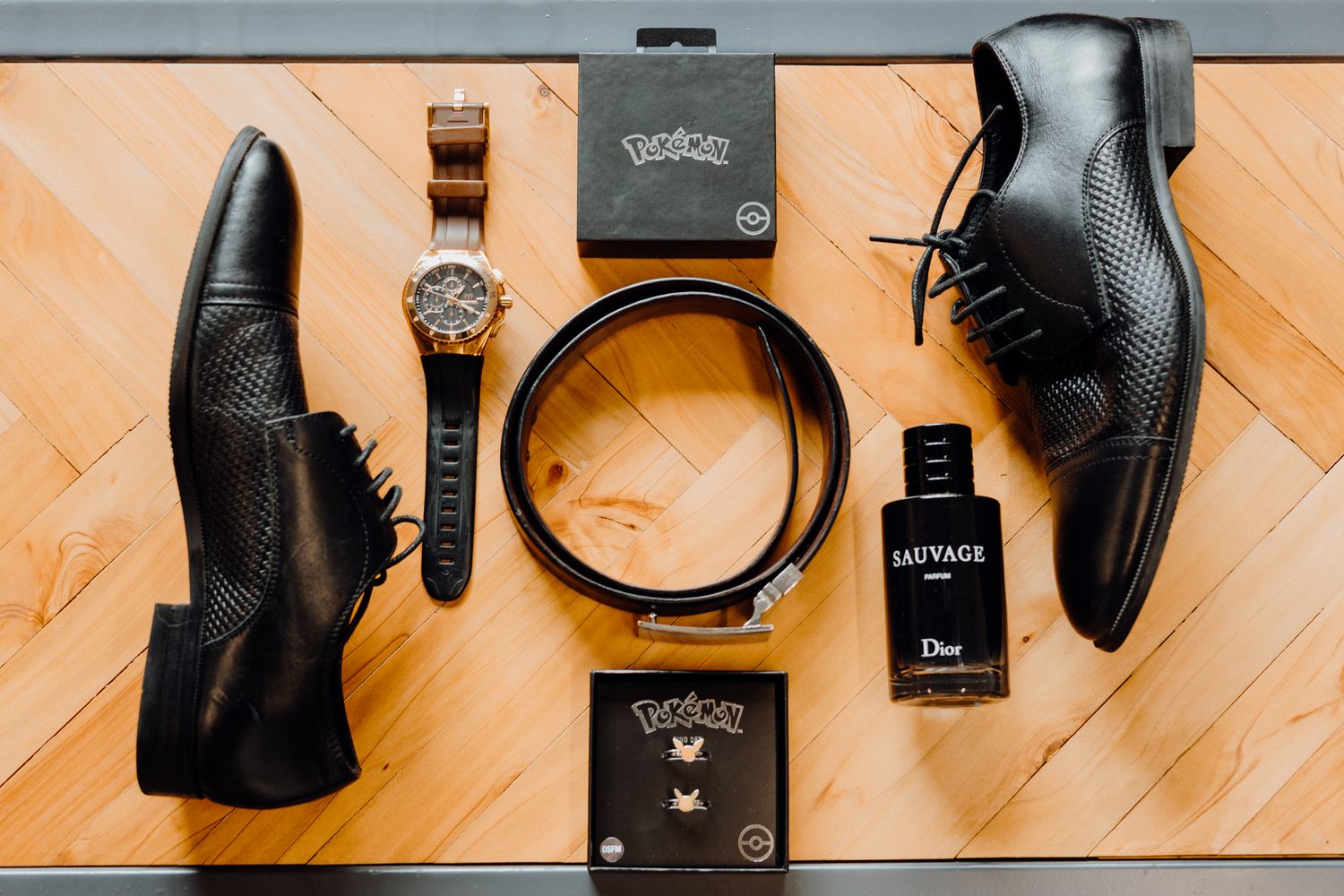 Wedding accessories including black dress shoes, belt, watch and perfume arranged on wooden floor.