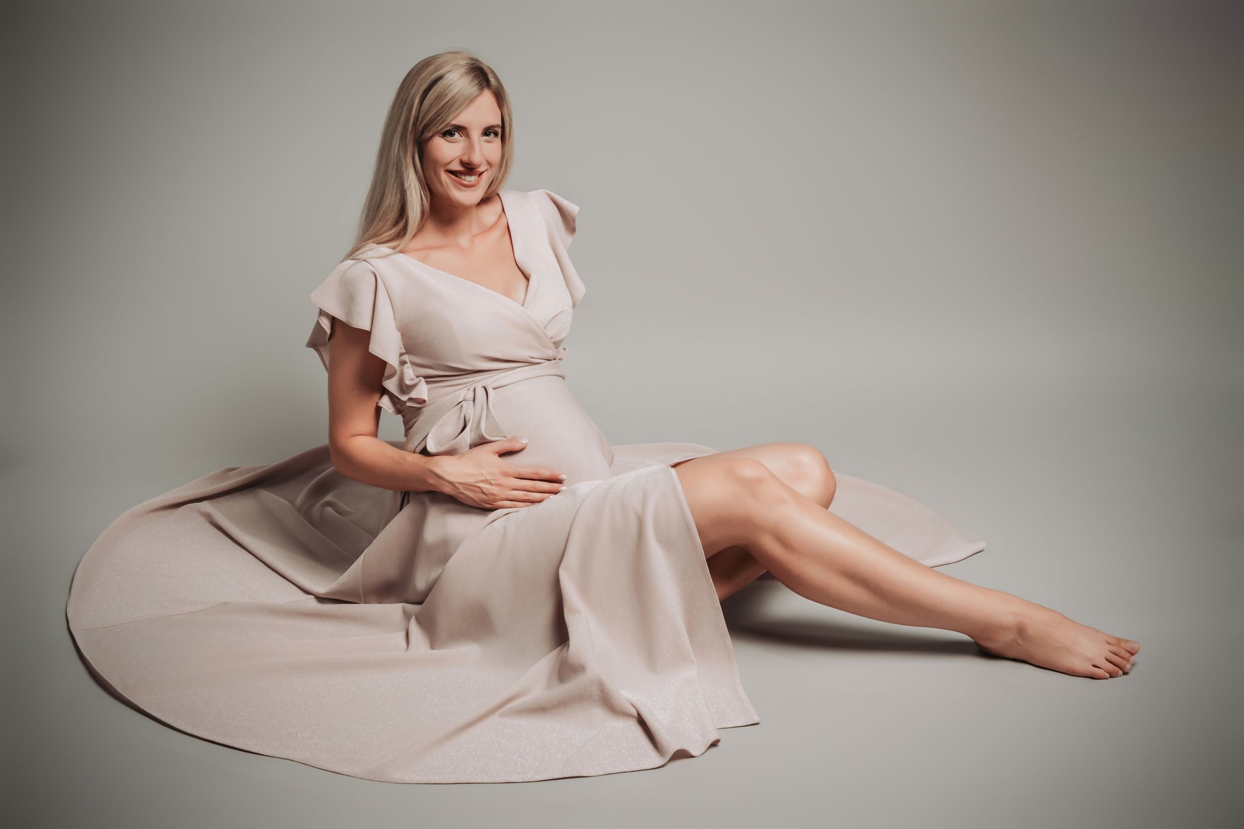 A maternity photo of a woman in a flowing beige dress on a grey background.