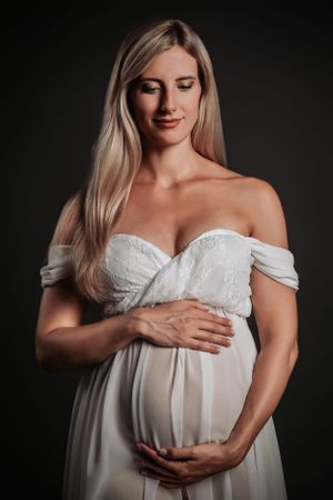 Maternity portrait of an expectant mother in white off-shoulder dress against dark backdrop.