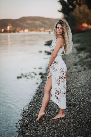 Woman in floral white dress poses on rocky shore at sunset with mountains in background.