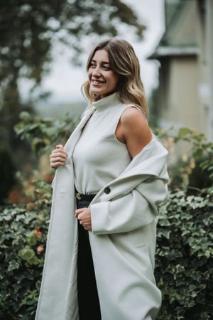 Woman in white sleeveless outfit and coat posing outdoors with greenery.