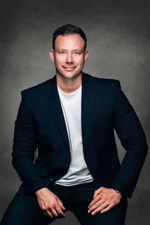 Professional headshot of man in navy blazer and white t-shirt against dark background.