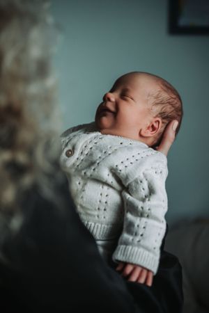 Smiling newborn in white knit sweater being held up.