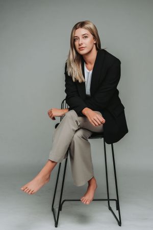 Professional studio portrait of woman in black blazer and khaki pants sitting barefoot on minimal stool.