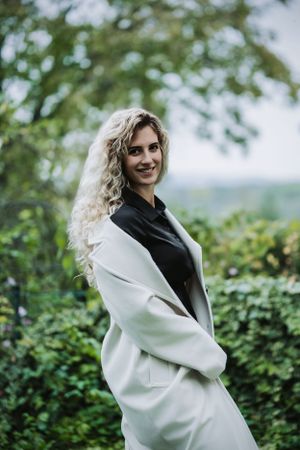 Portrait of woman in white coat smiling against green foliage background.