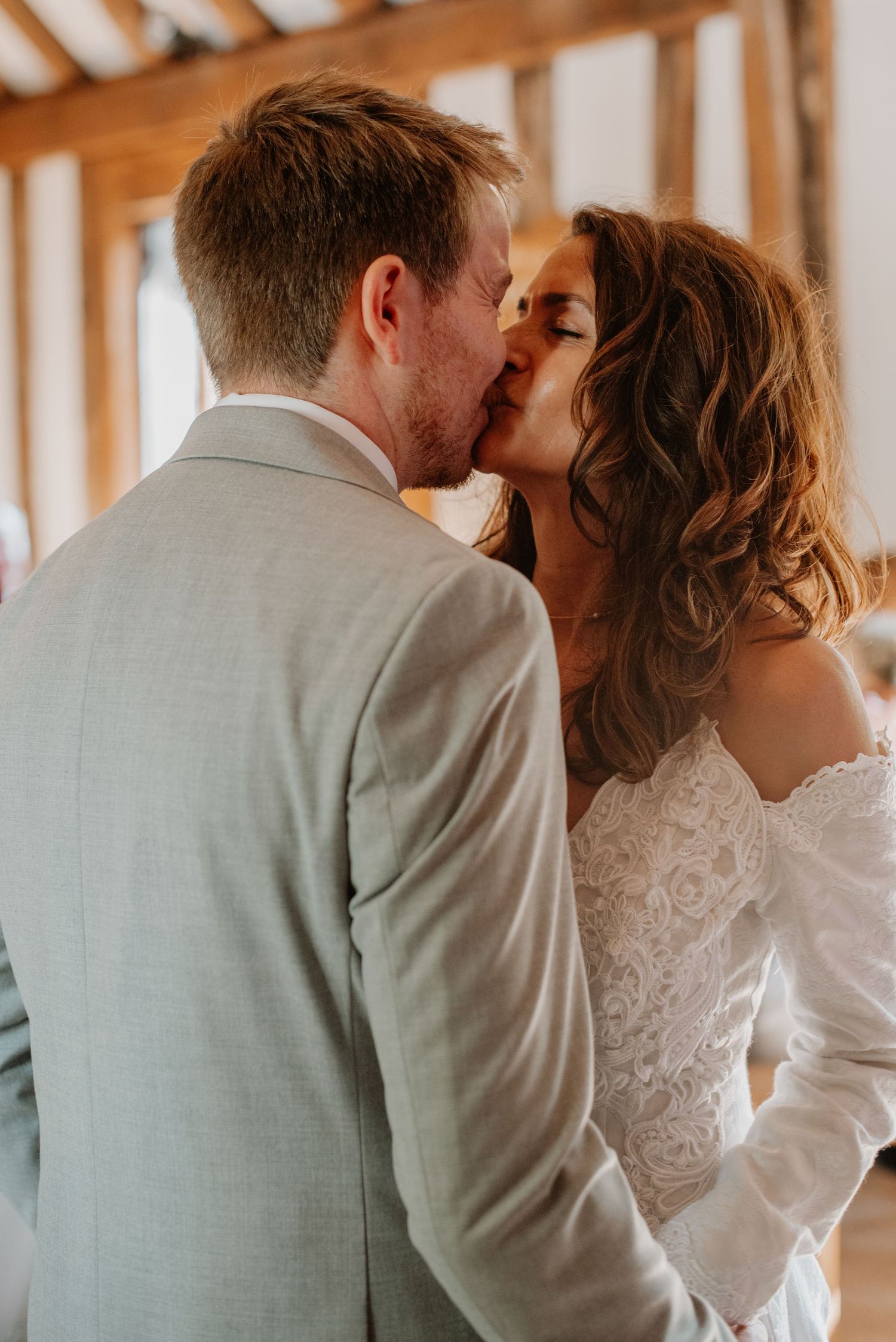 A romantic kiss between newlyweds in a rustic indoor setting, with soft lighting highlighting the bride's lace dress.