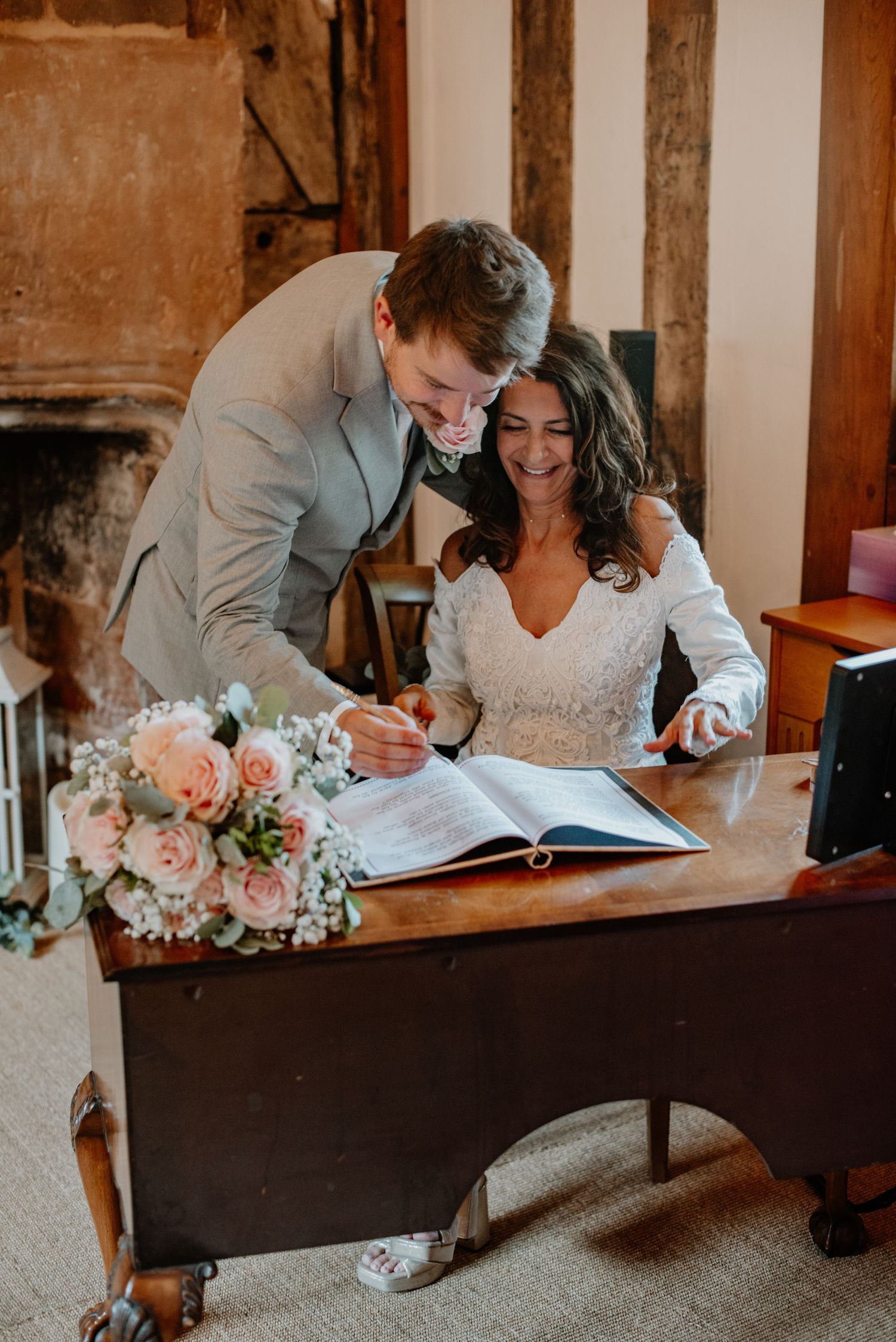 A couple in wedding attire signing documents together while smiling, with a pink and white floral bouquet on the desk.