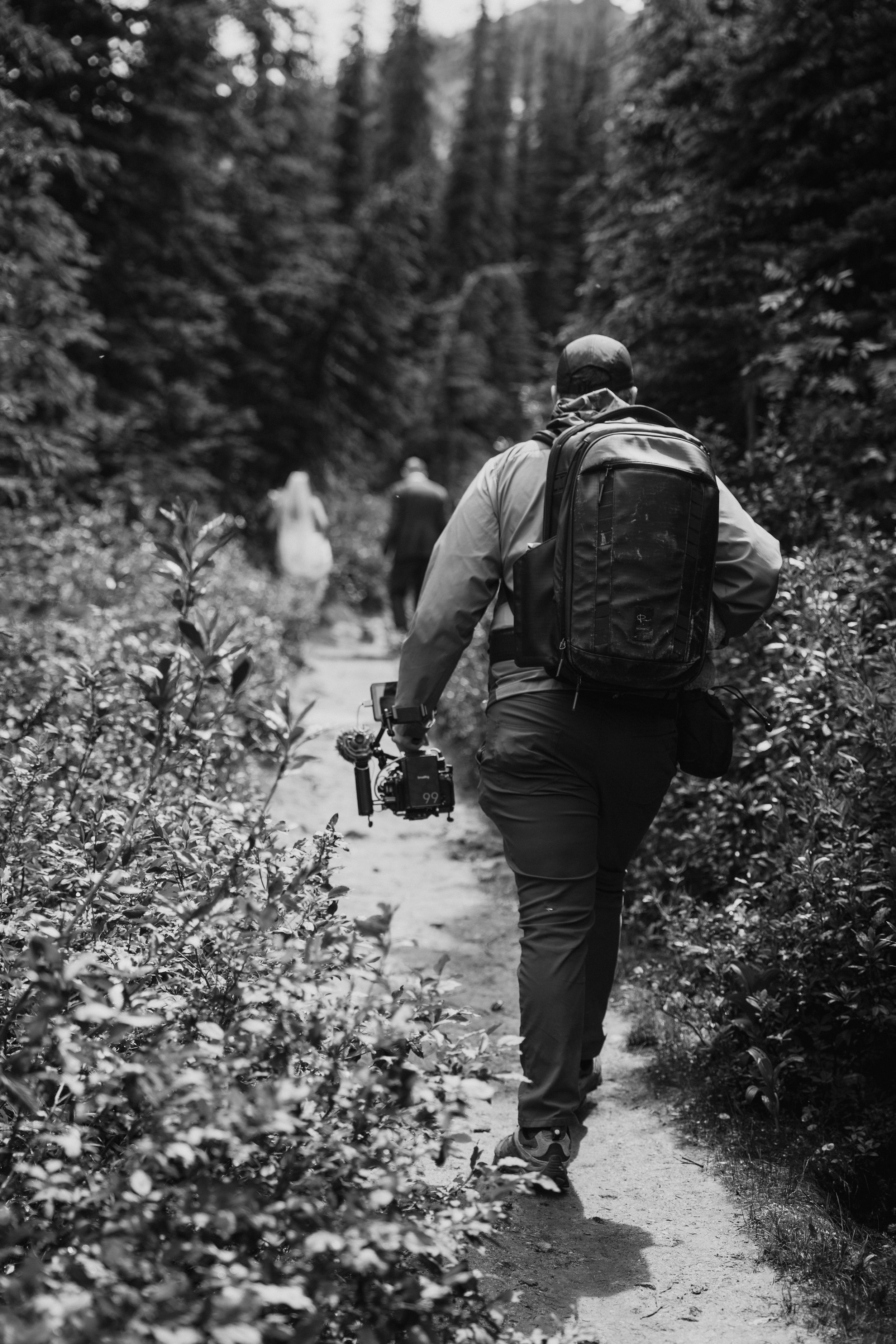 Hikers walk along a forested mountain trail carrying backpacks through evergreen wilderness in black and white.