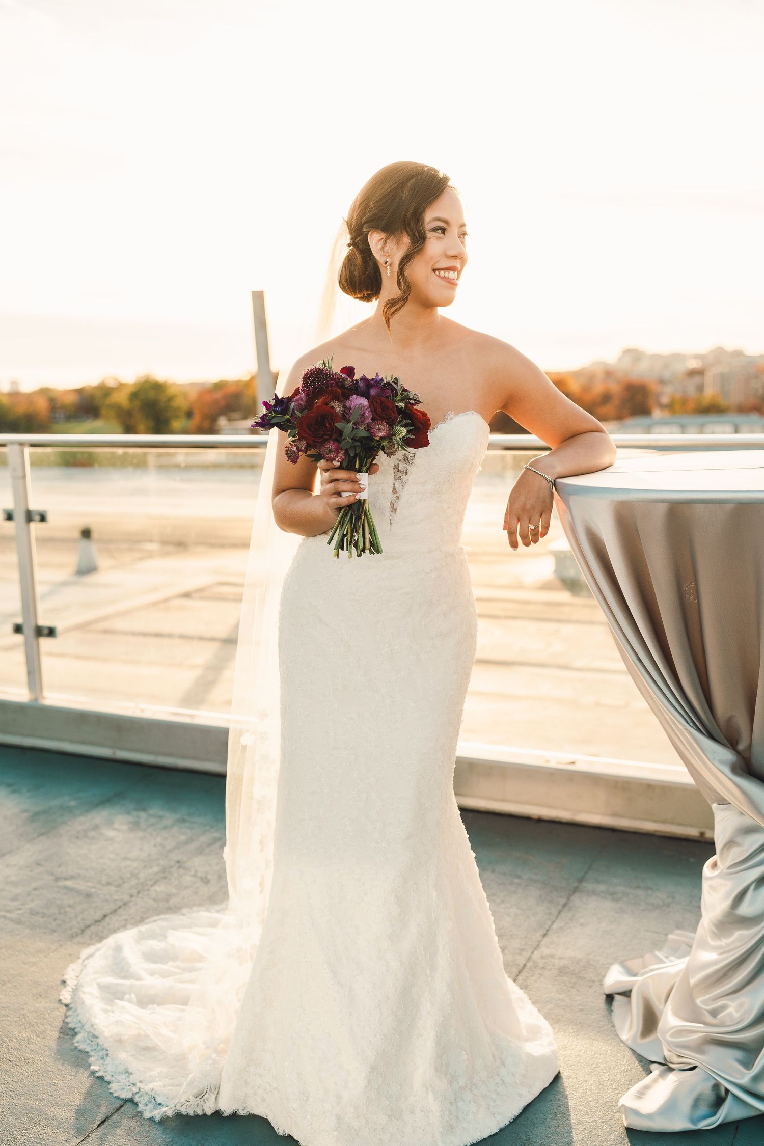 A bride in a fitted white strapless gown holds a deep purple bouquet on a rooftop terrace at sunset.