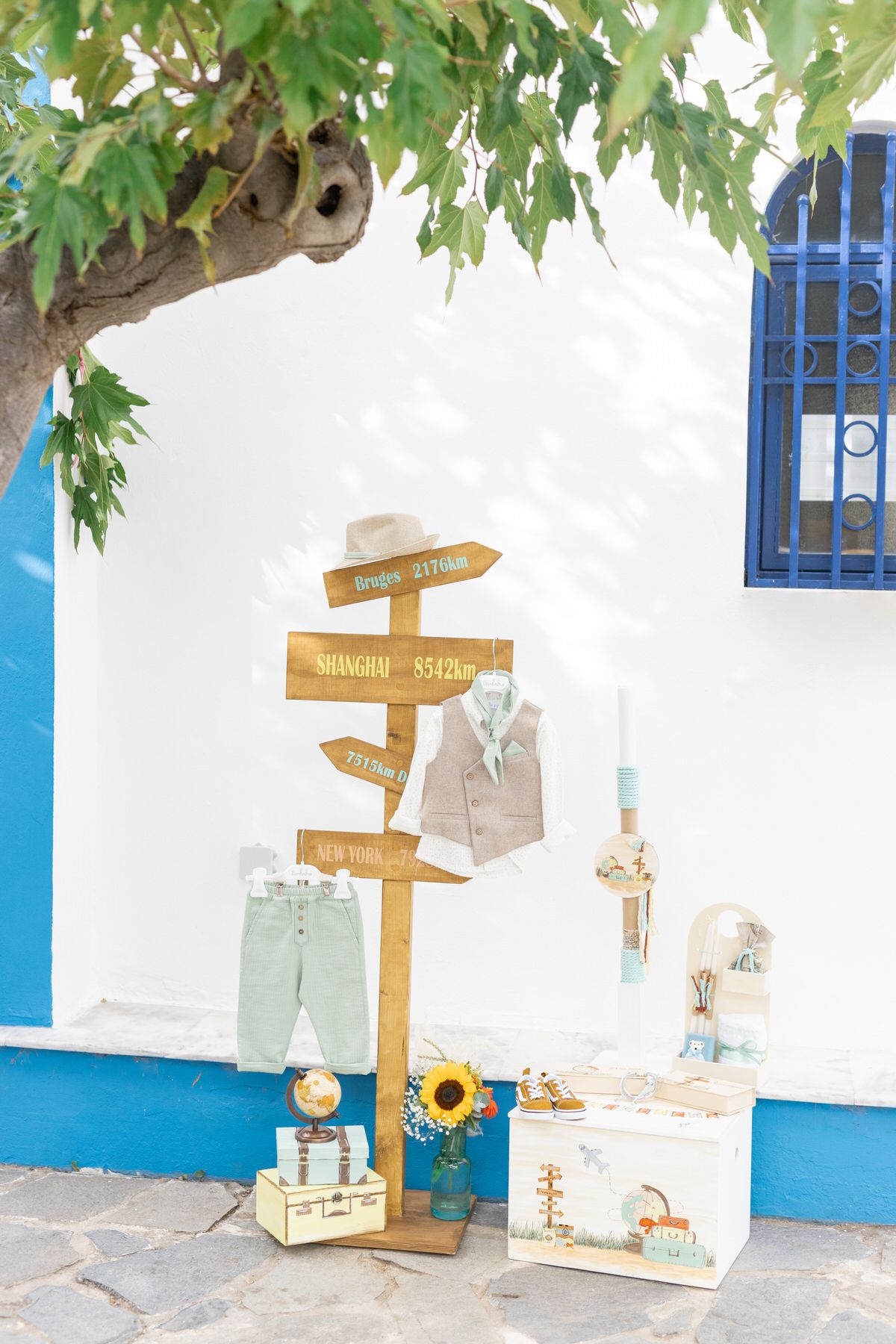 A rustic wooden directional sign post with vintage clothes and decor against a white wall with blue trim and green foliage.