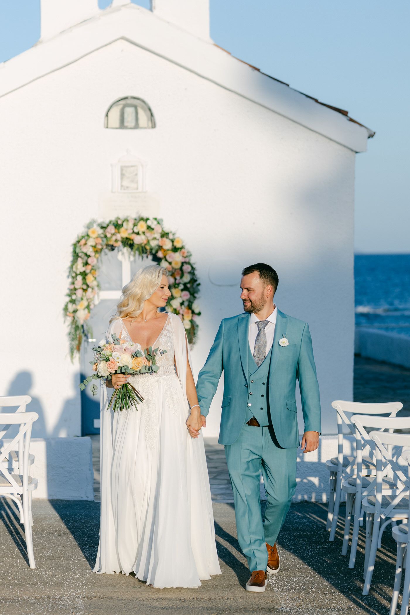 Newlyweds walk hand in hand by a white chapel along the sea under a floral arch in Greece.
