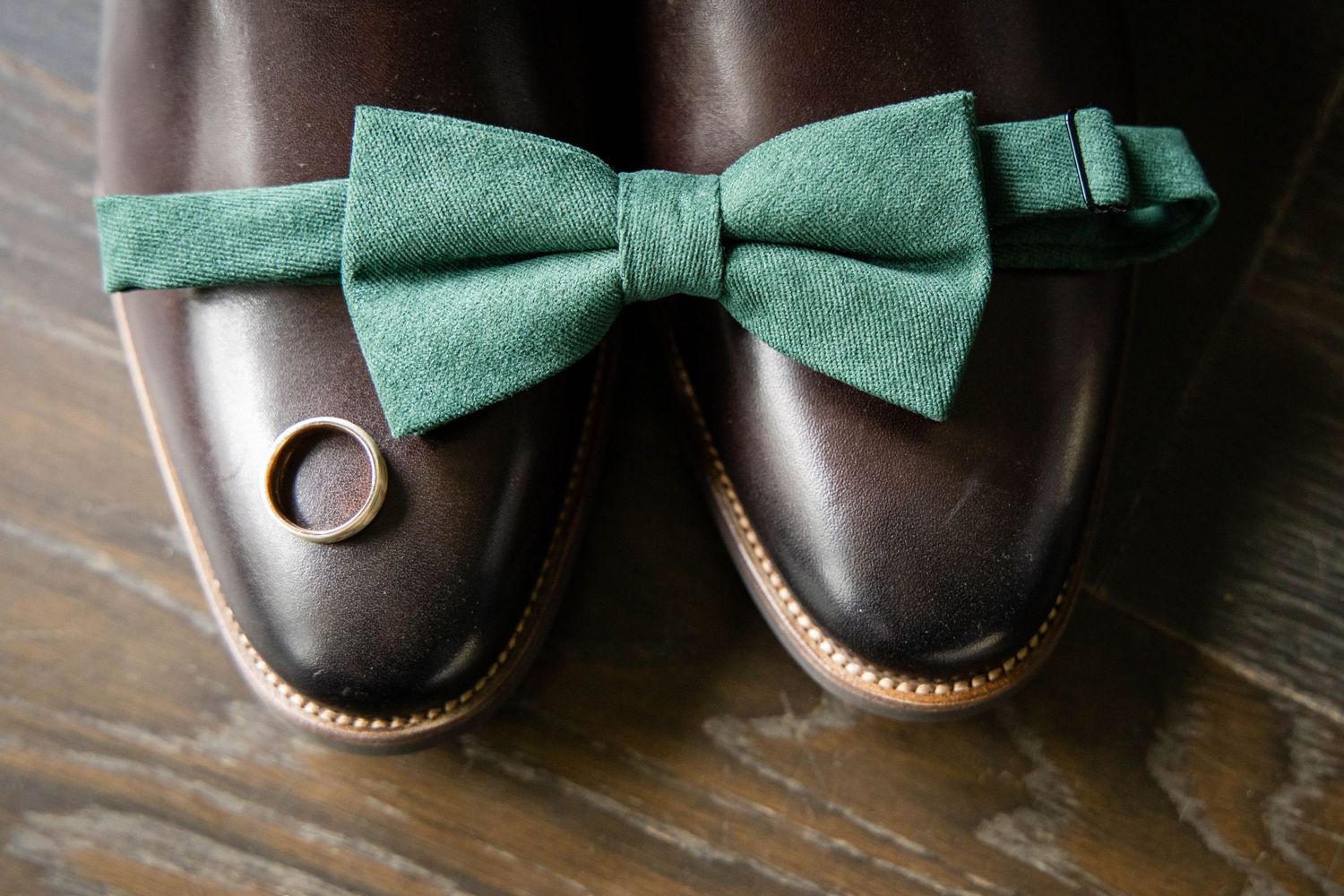 A pair of brown leather dress shoes with a green bow tie placed across them on a wooden surface.