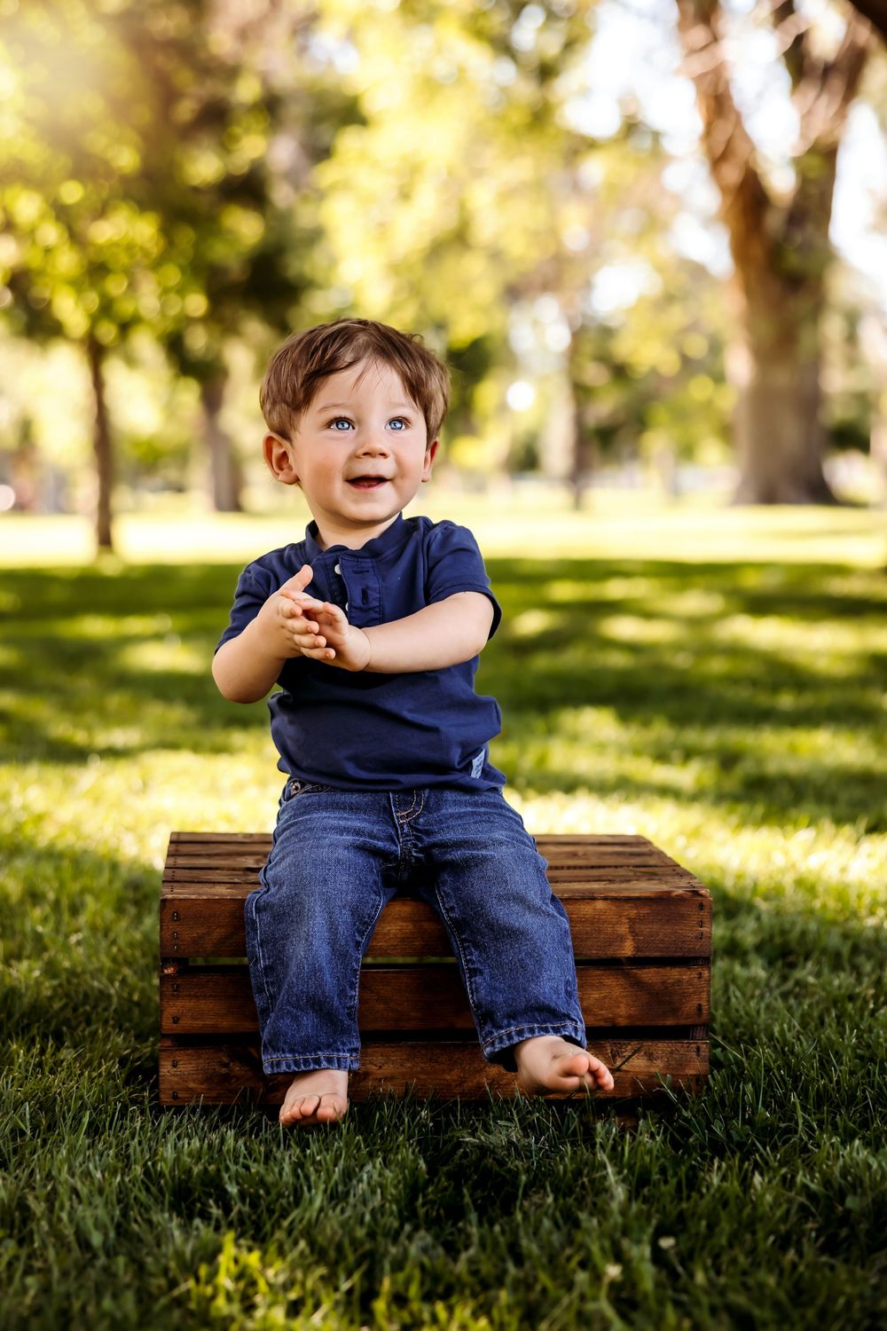 Child wearing navy blue shirt and jeans sits barefoot on wooden crate in sunny park with bokeh background. Pueblo Photographer