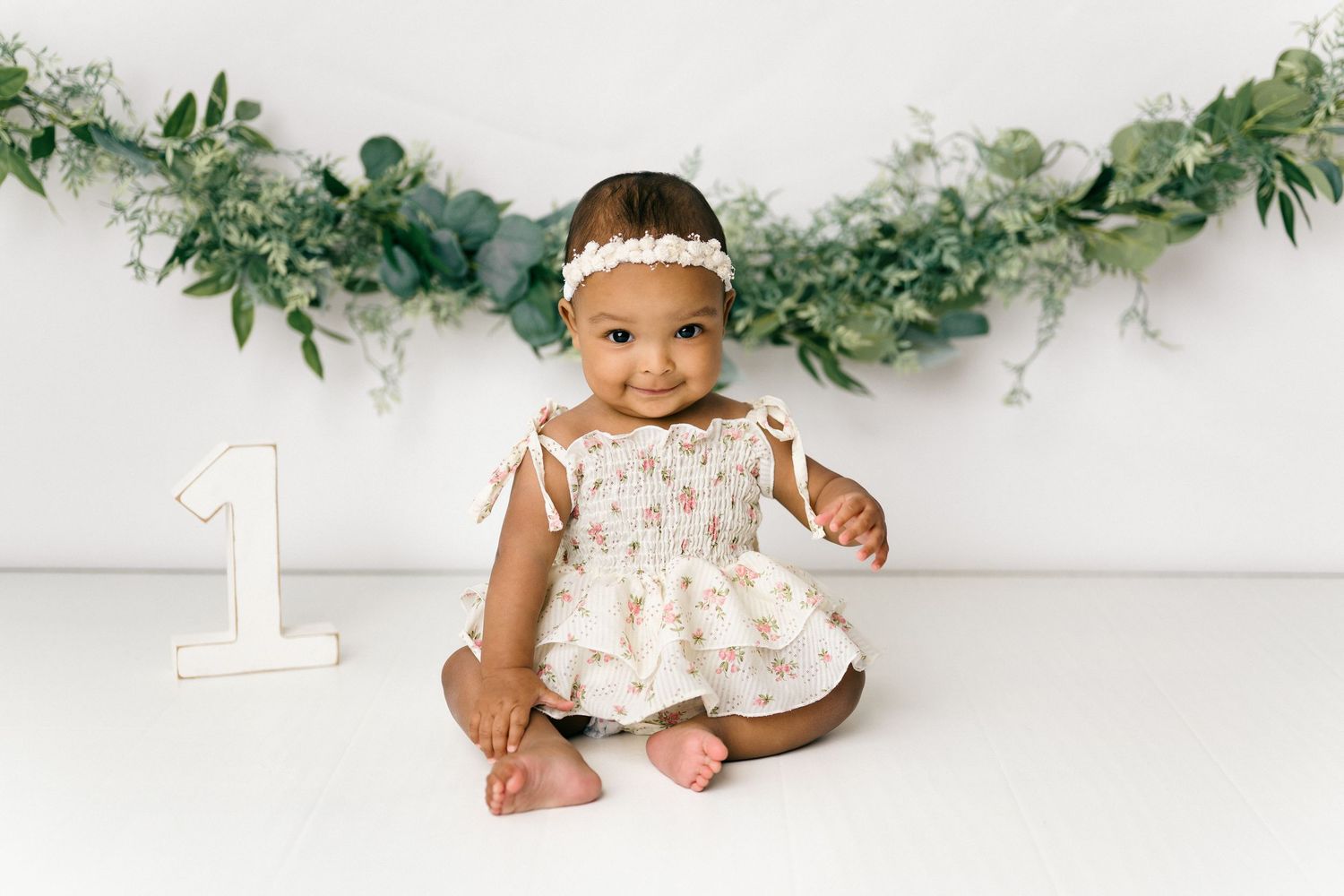 A toddler in a pink tulle dress sits on a white floor with a white cake, beneath floral decor.