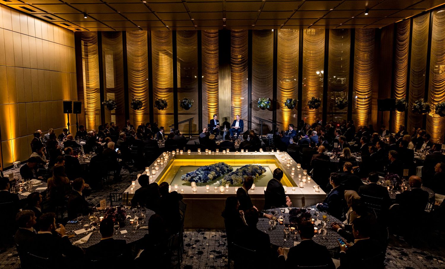 An elegant banquet hall with a brightly lit center stage surrounded by round dining tables in a dimly lit atmosphere.