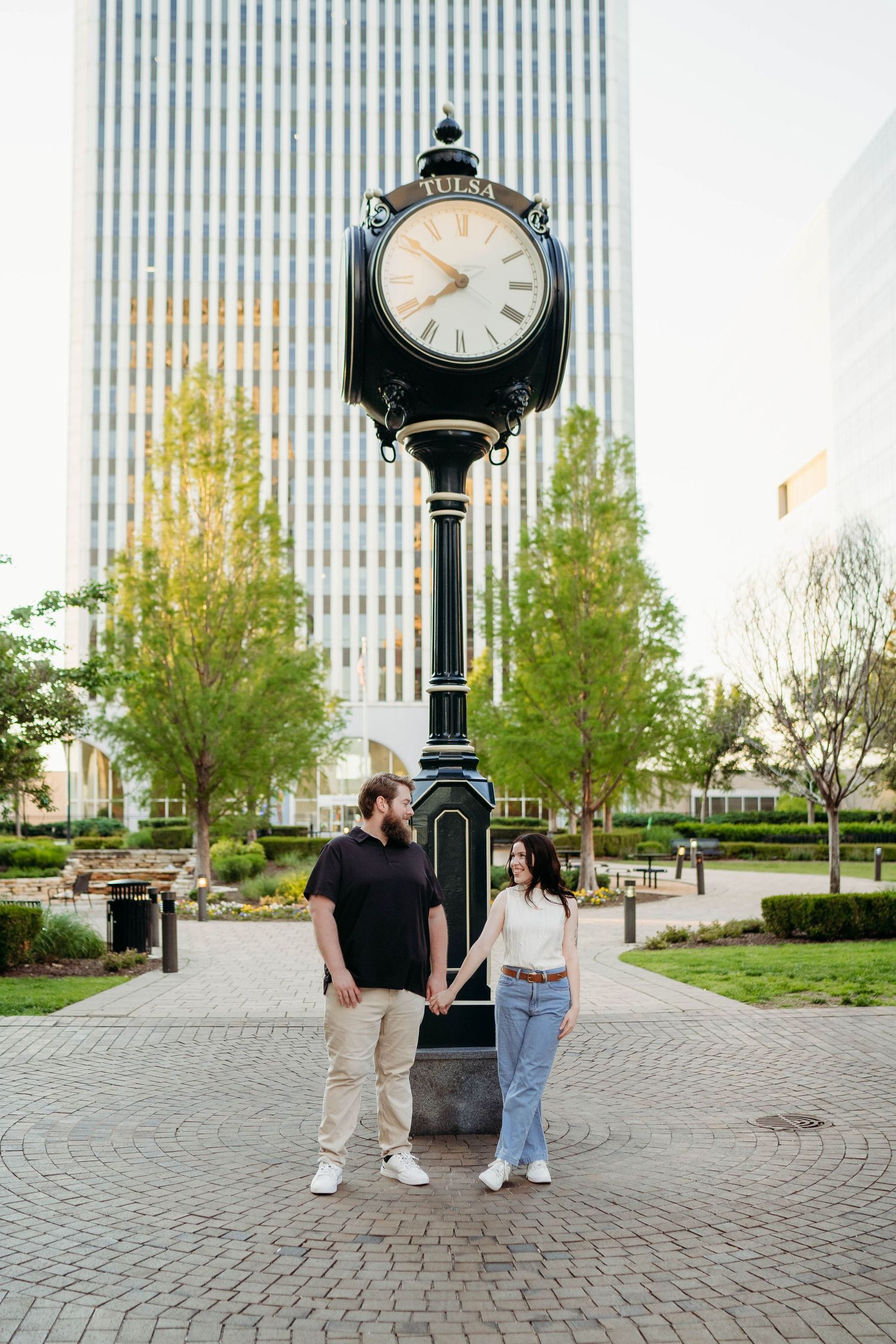 Two people stand under a vintage-style clock tower on a brick paved plaza with trees and buildings in the background.