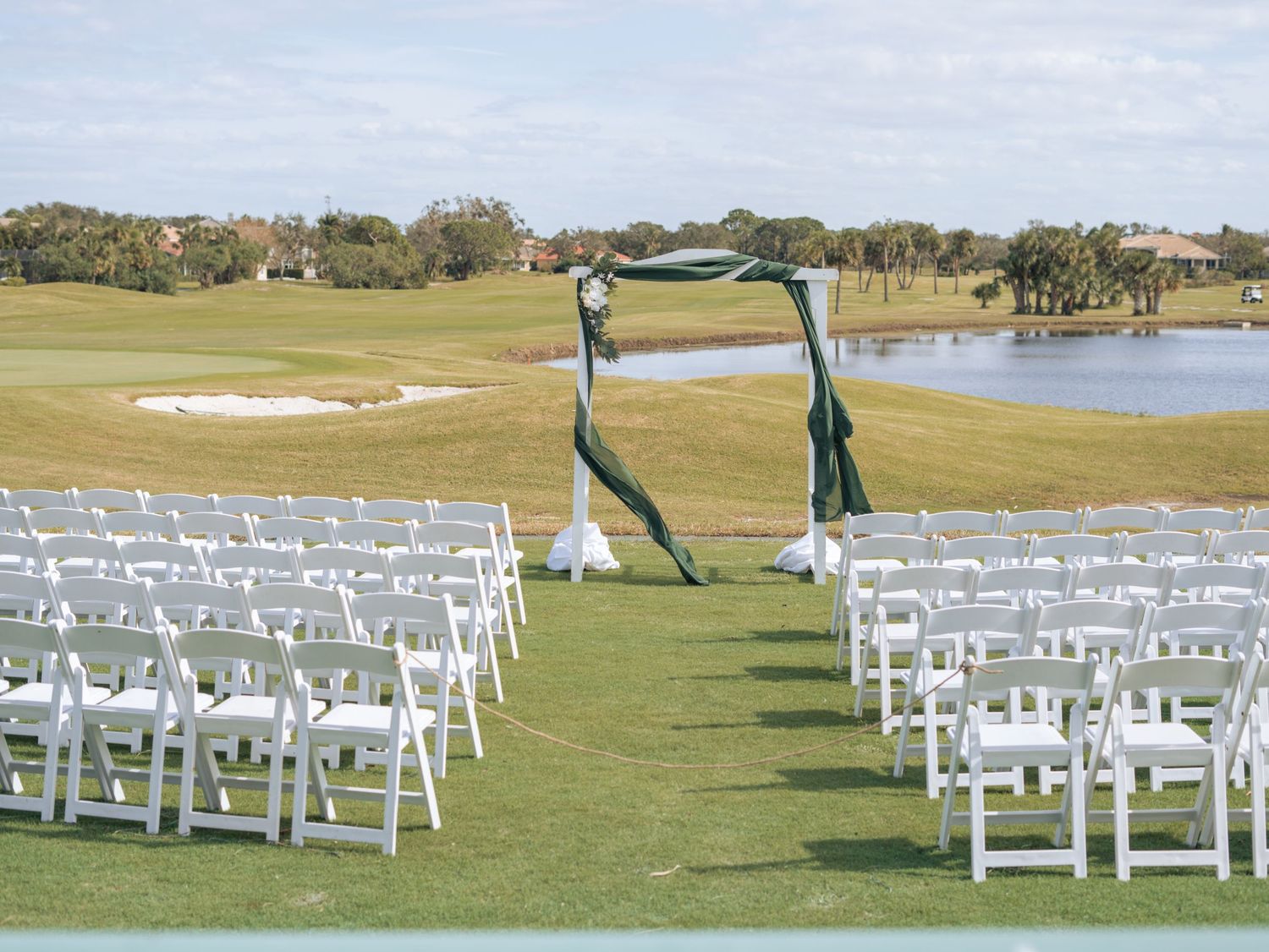 Wedding ceremony setup on a golf course with white chairs and a green archway overlooking a pond.