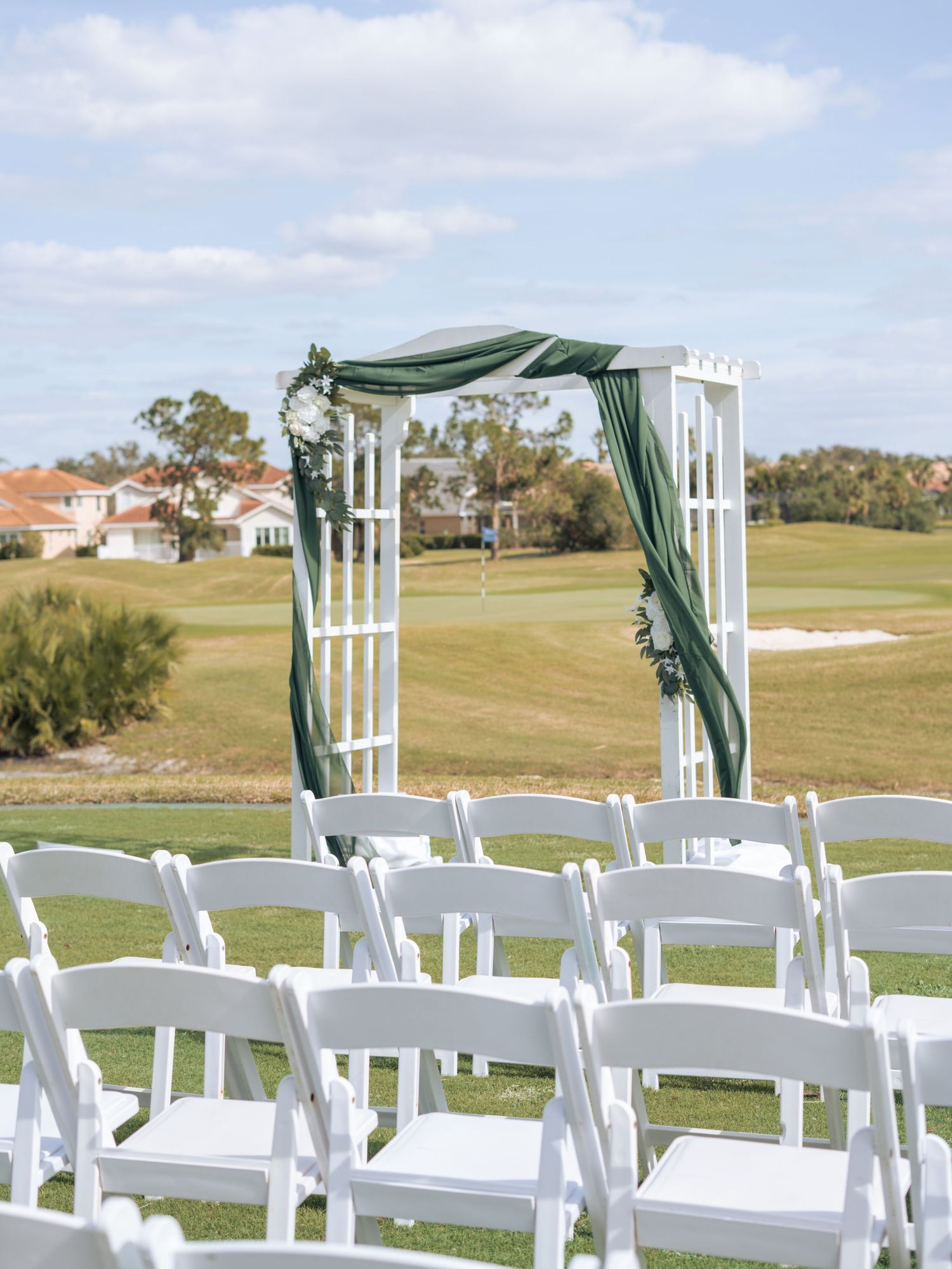 White wedding ceremony setup with draped arch and chairs on a golf course under blue sky.