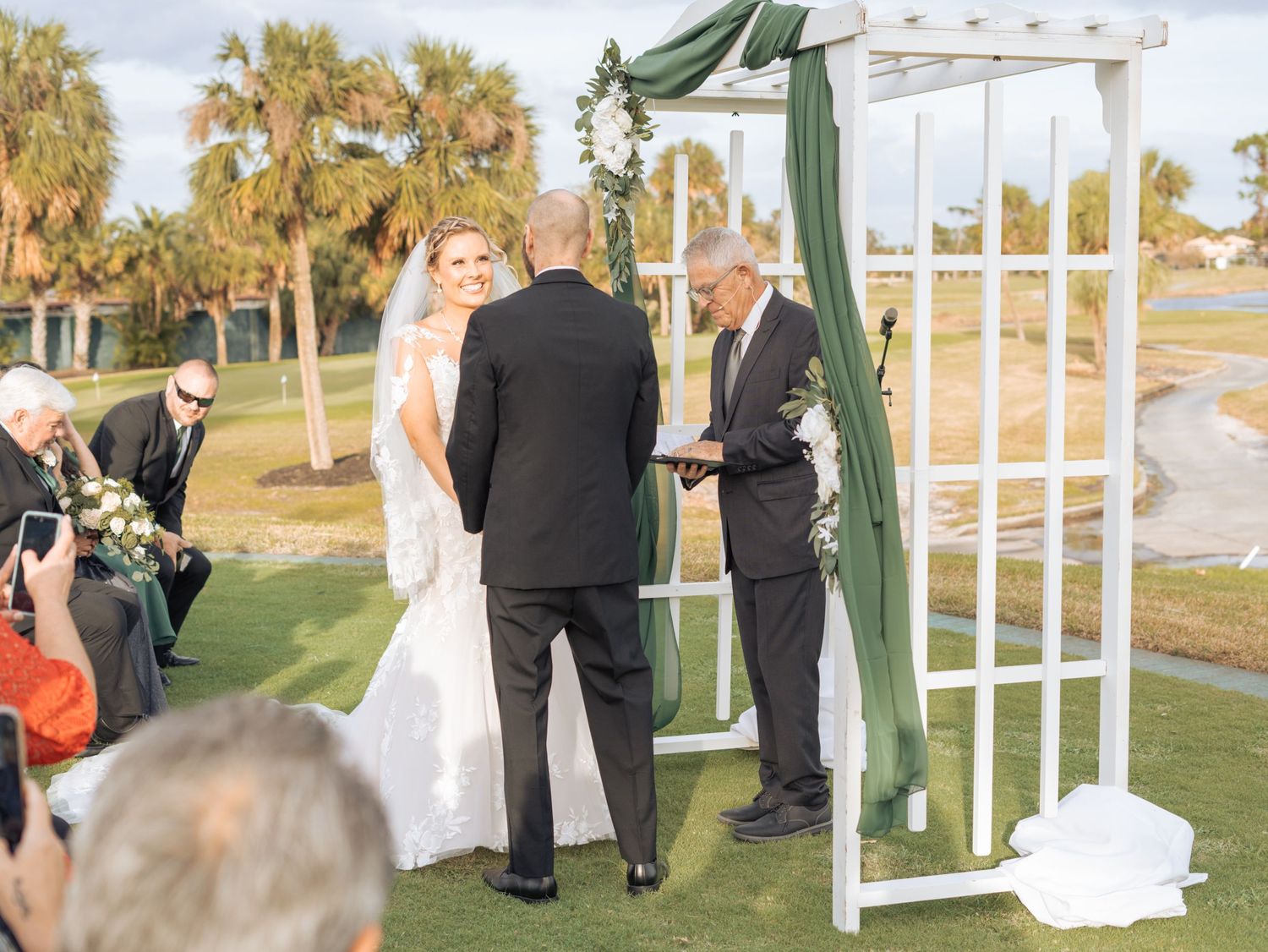 Wedding ceremony taking place under a white pergola decorated with green fabric on a golf course.