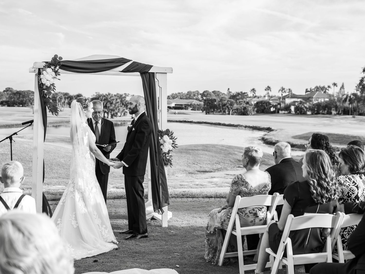 Black and white photo of an outdoor wedding ceremony with guests seated in white chairs.