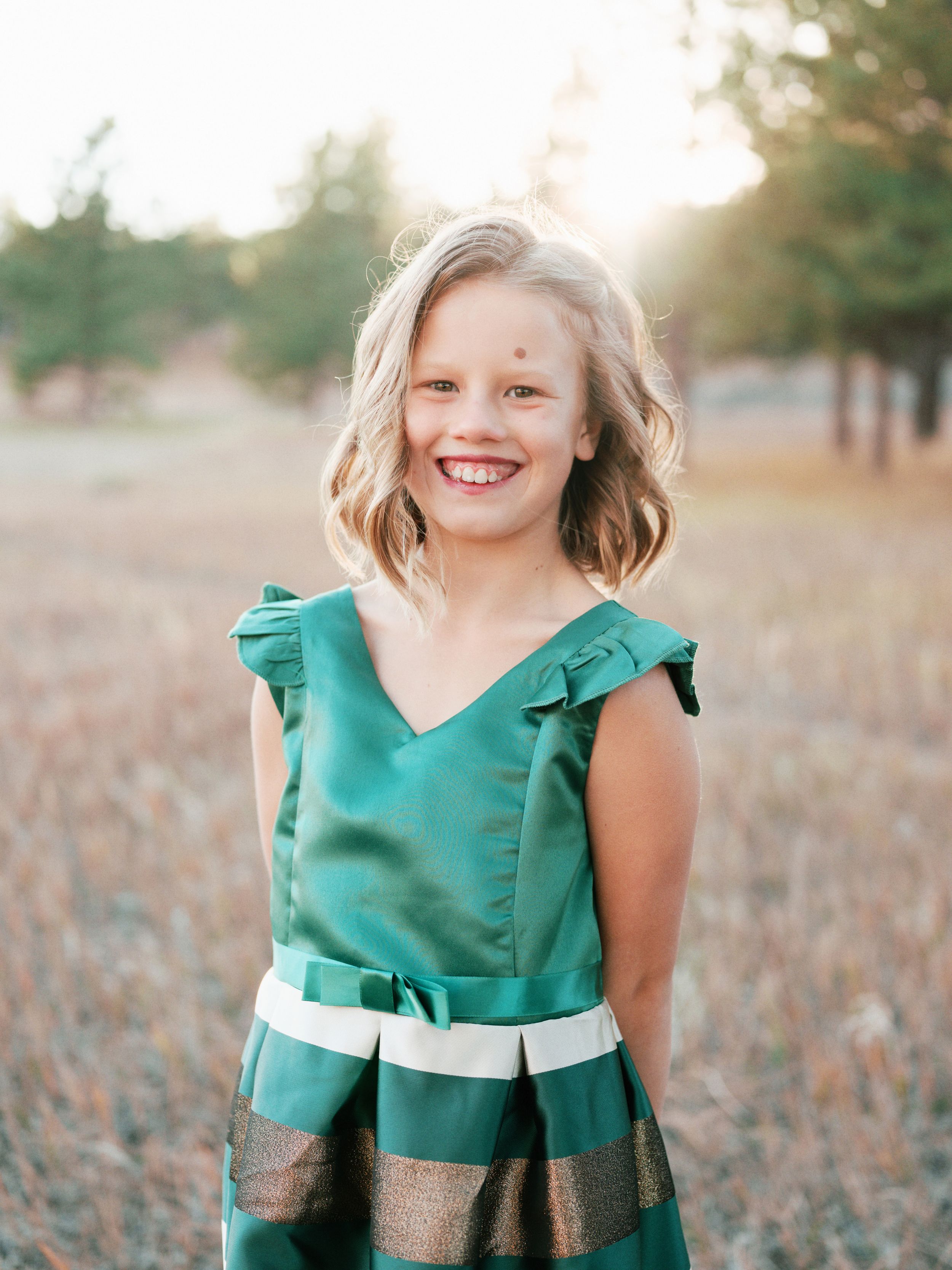 Young person wearing a green dress with ruffled sleeves and striped skirt standing in a field at sunset.