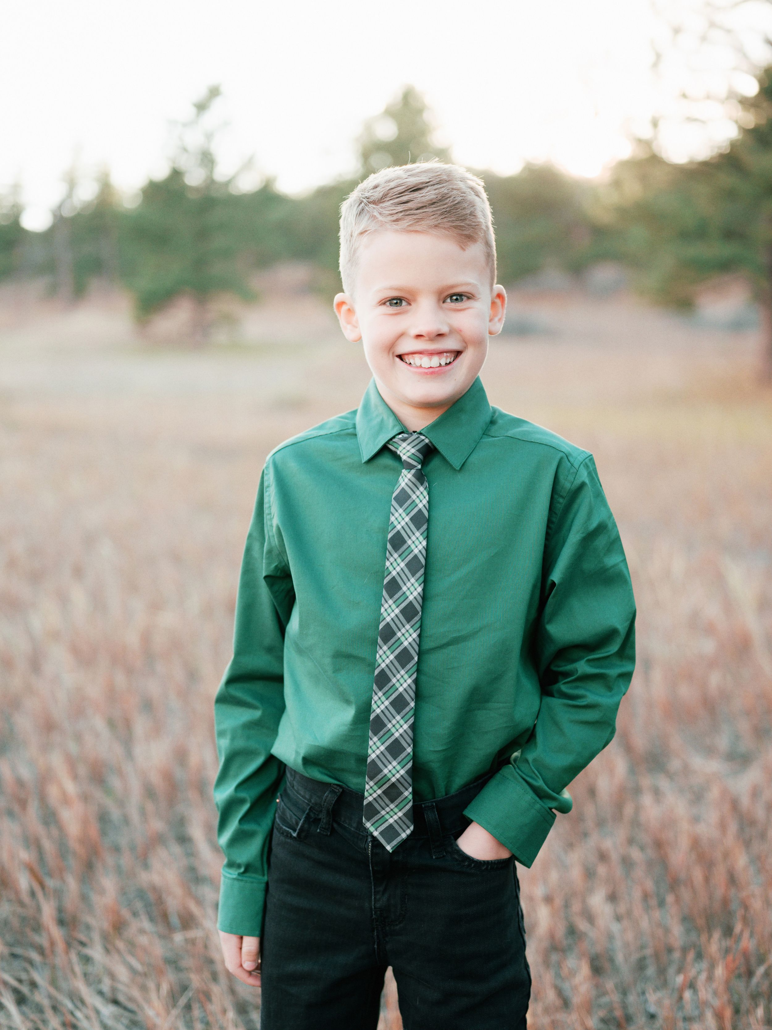 Child in green dress shirt and plaid tie smiling while standing in outdoor field setting.