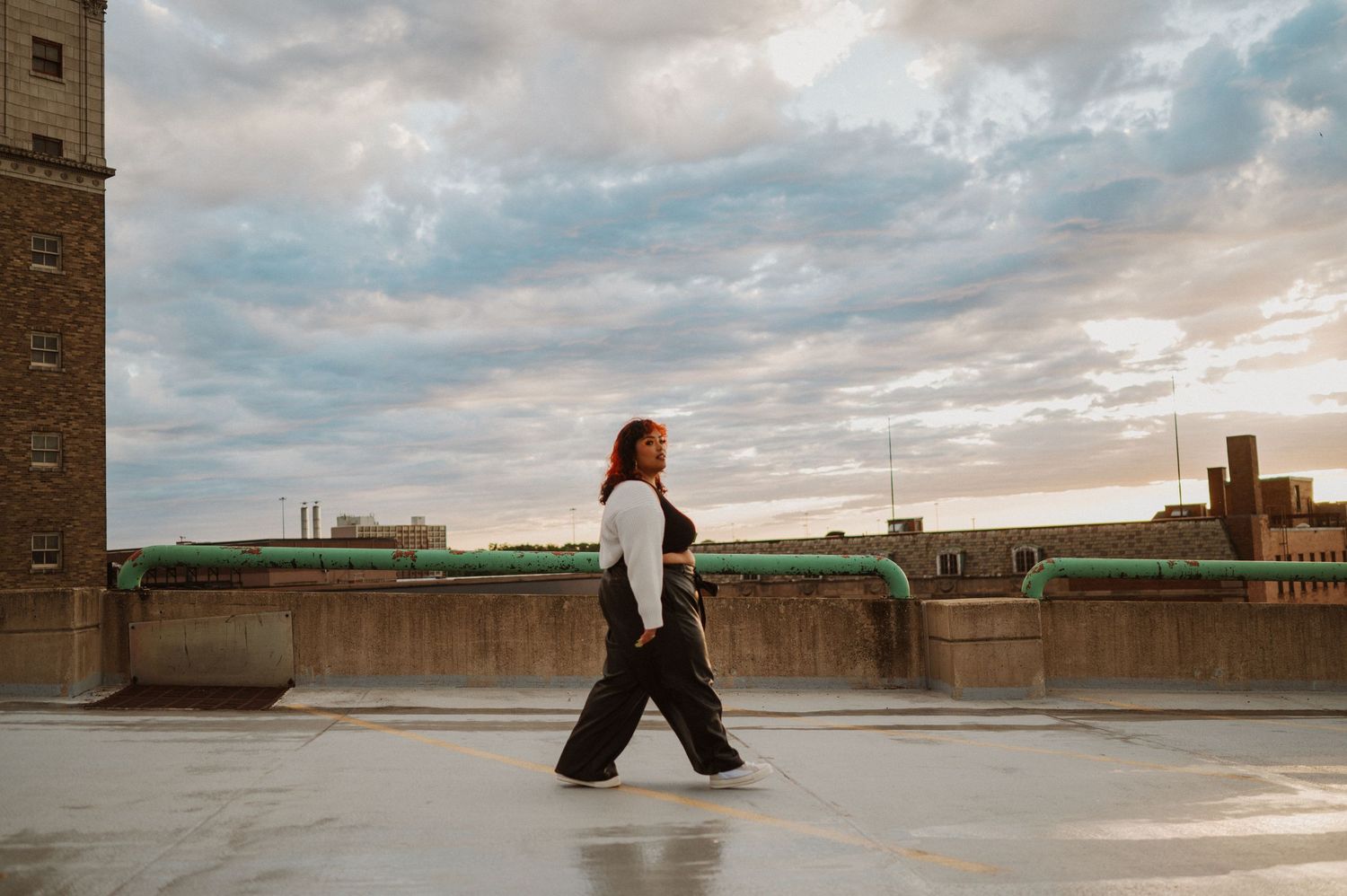Person in dark pants and white shirt walks across an urban rooftop terrace under dramatic cloudy sky at sunset.
