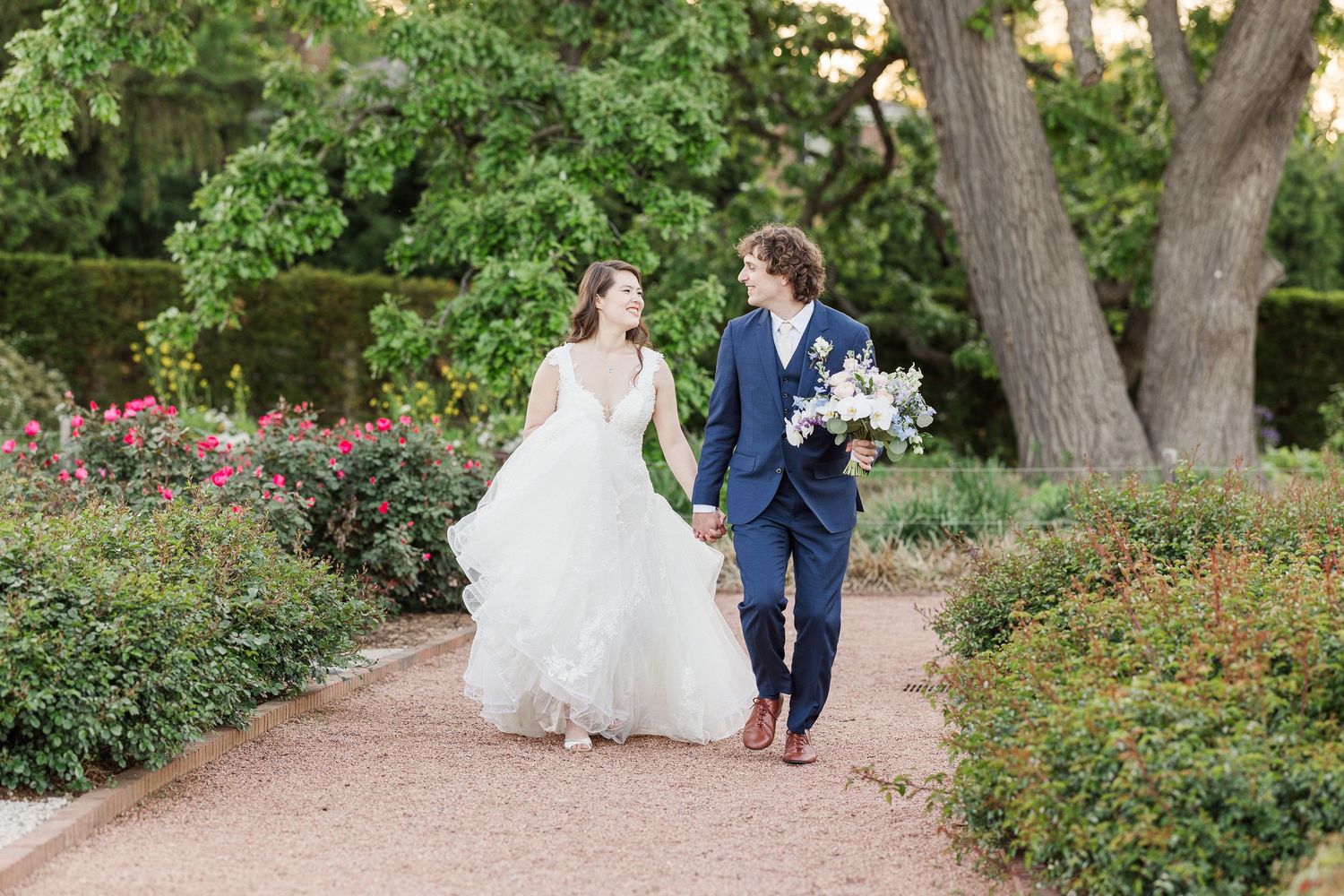 A couple walks together along a garden path surrounded by lush greenery and blooming flowers on their wedding day.