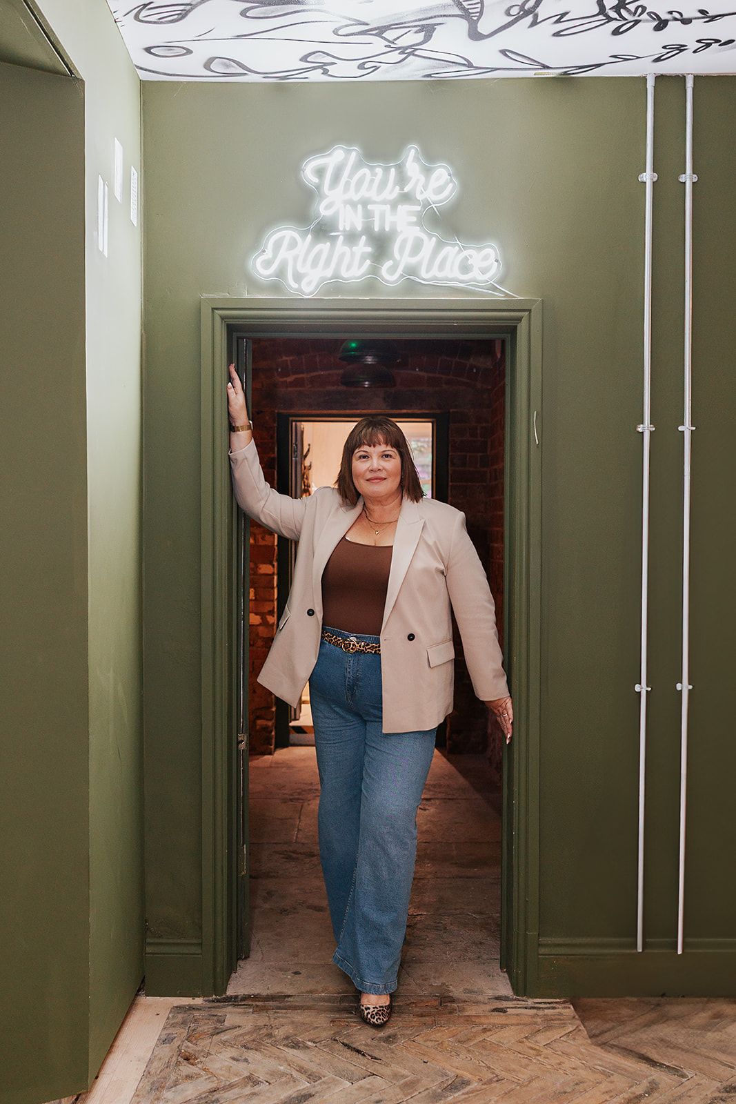Someone stands in a doorway beneath a neon sign reading 'You're in the Right Place' wearing jeans and a beige blazer.