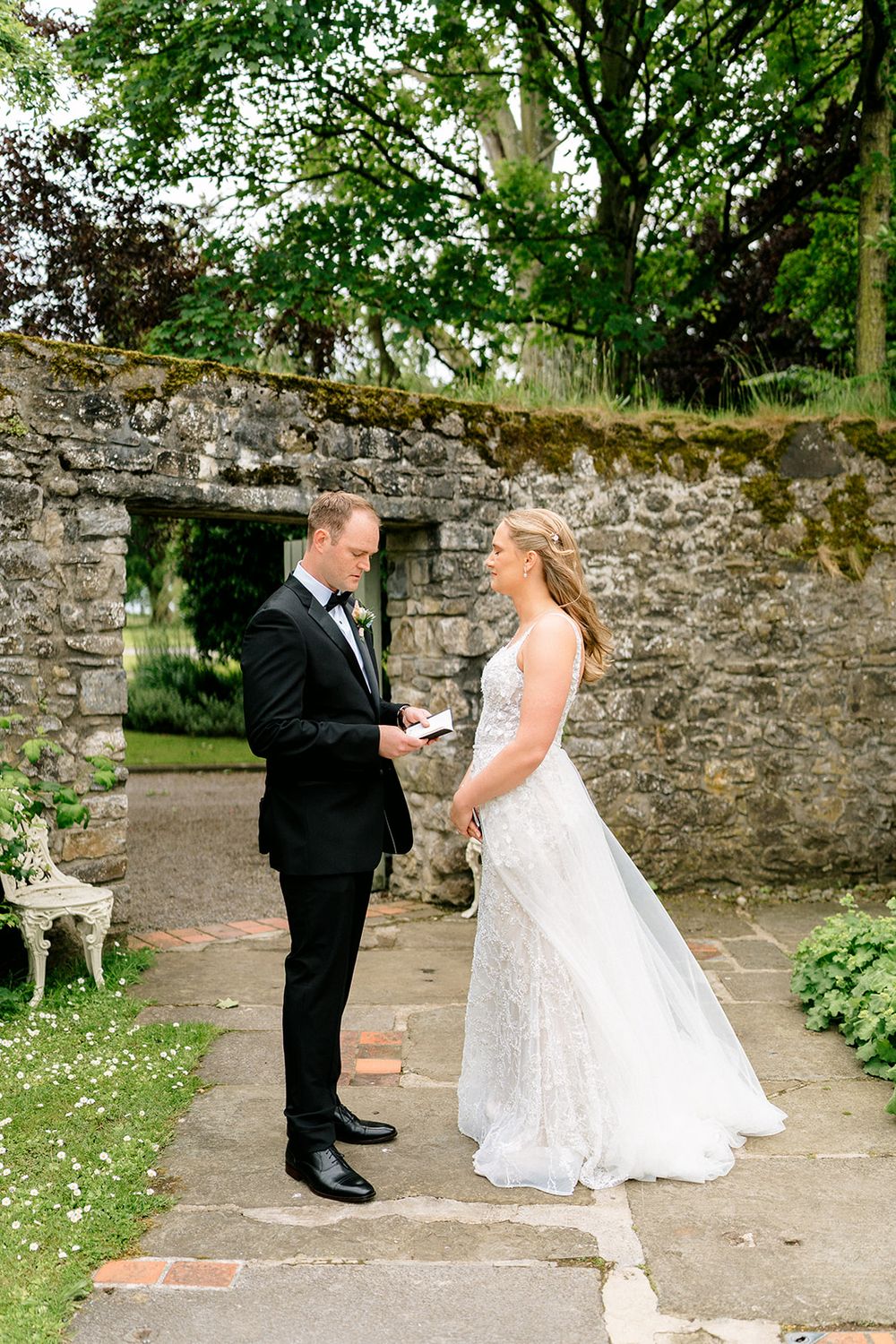 Two people in wedding attire share a sweet moment in front of an old stone wall during their first look photo session.