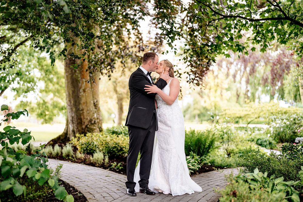 Couple embraces on stone pathway under lush green trees in romantic garden wedding setting.