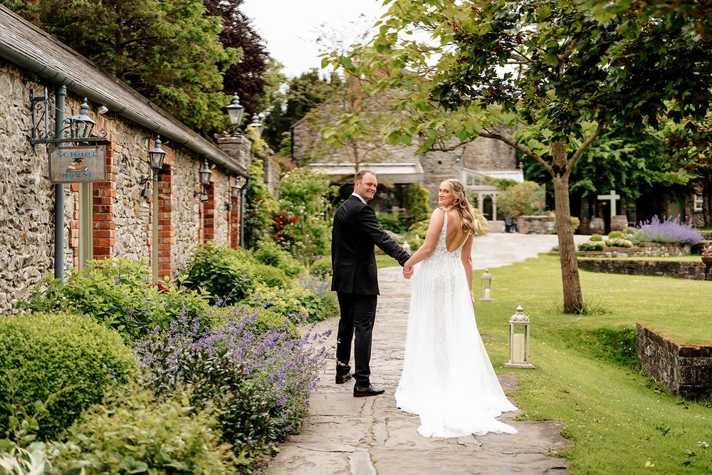 Wedding couple walking down tree-lined path at elegant outdoor venue with stone walls and lush gardens.