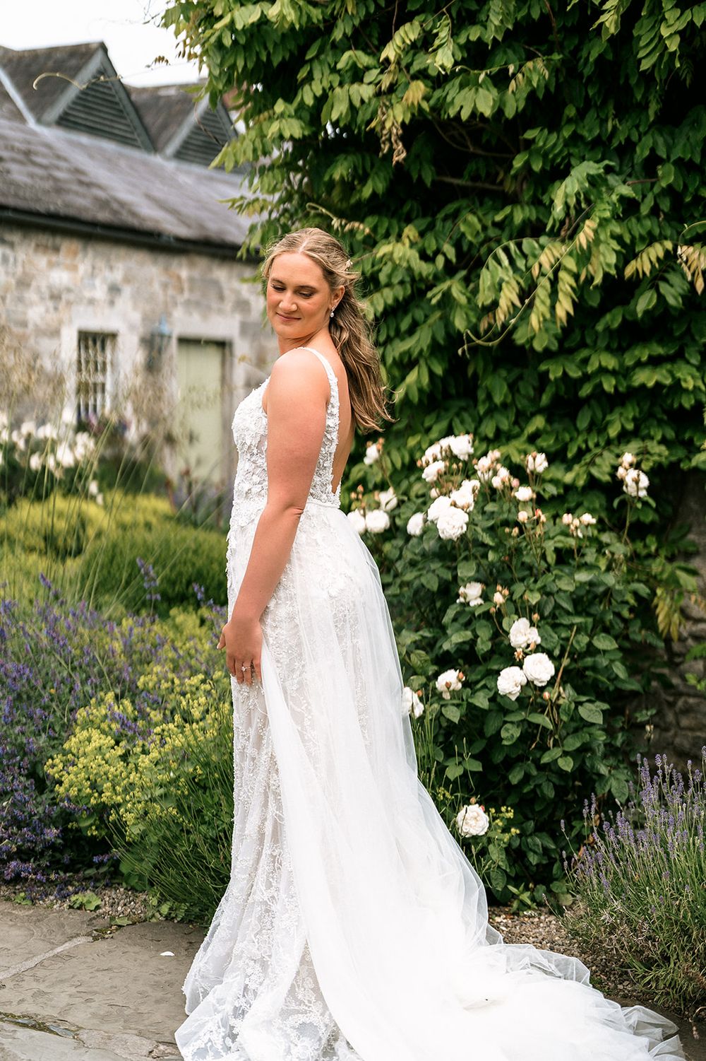 A bride in a white lace wedding dress with button detail poses in a garden setting with white roses and lavender.