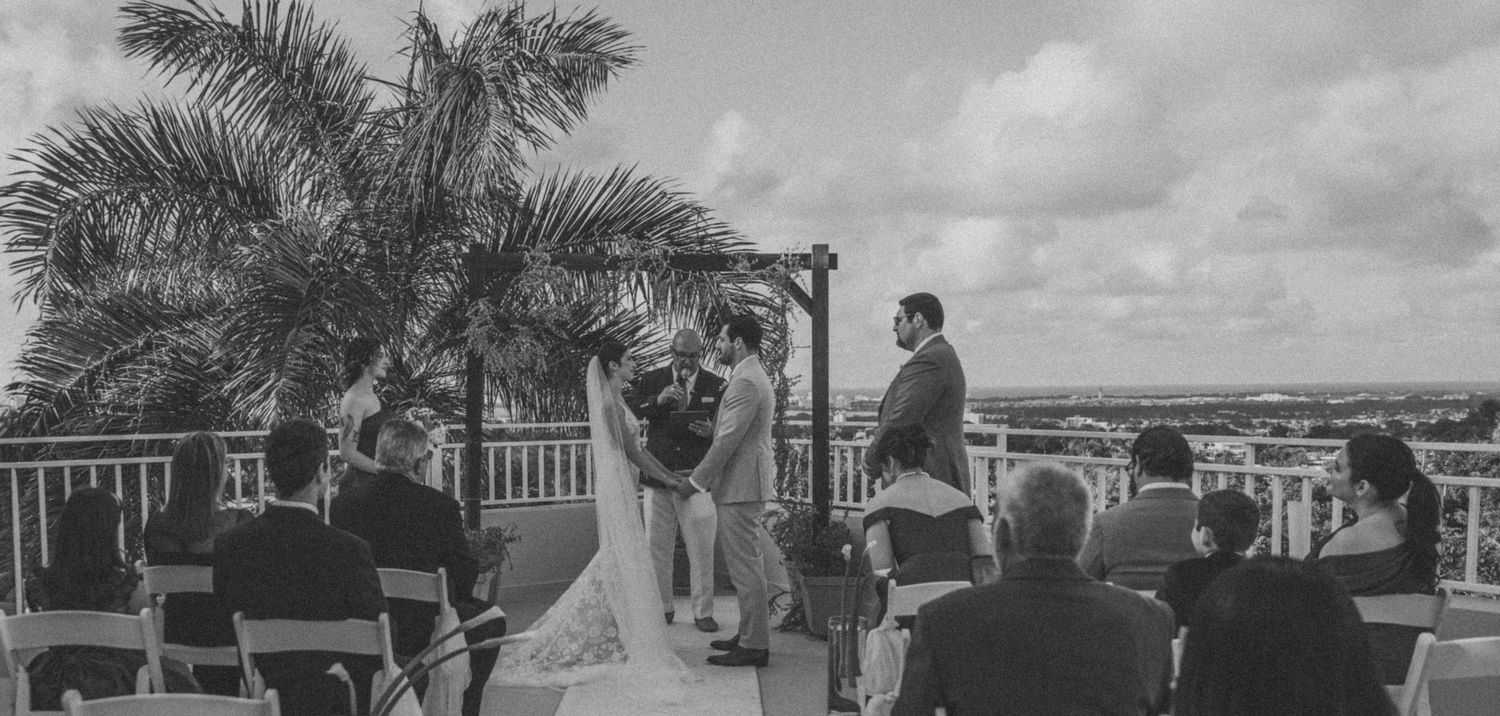 An intimate outdoor wedding ceremony at a covered pavilion overlooking scenic coastal views in black and white.