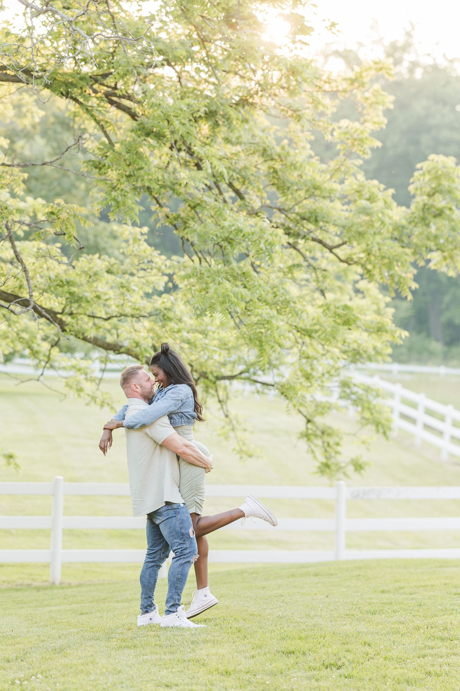A romantic couple embrace in a joyful moment on a grassy hill with white fencing and blooming trees in the background.
